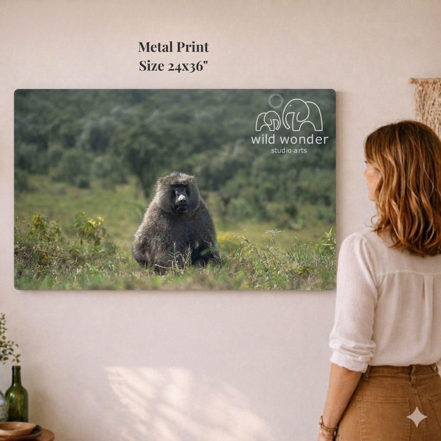 A woman is standing in front of a large metal print depicting a baboon in a grassy field, with the text "wild wonder studio arts" visible on the print.