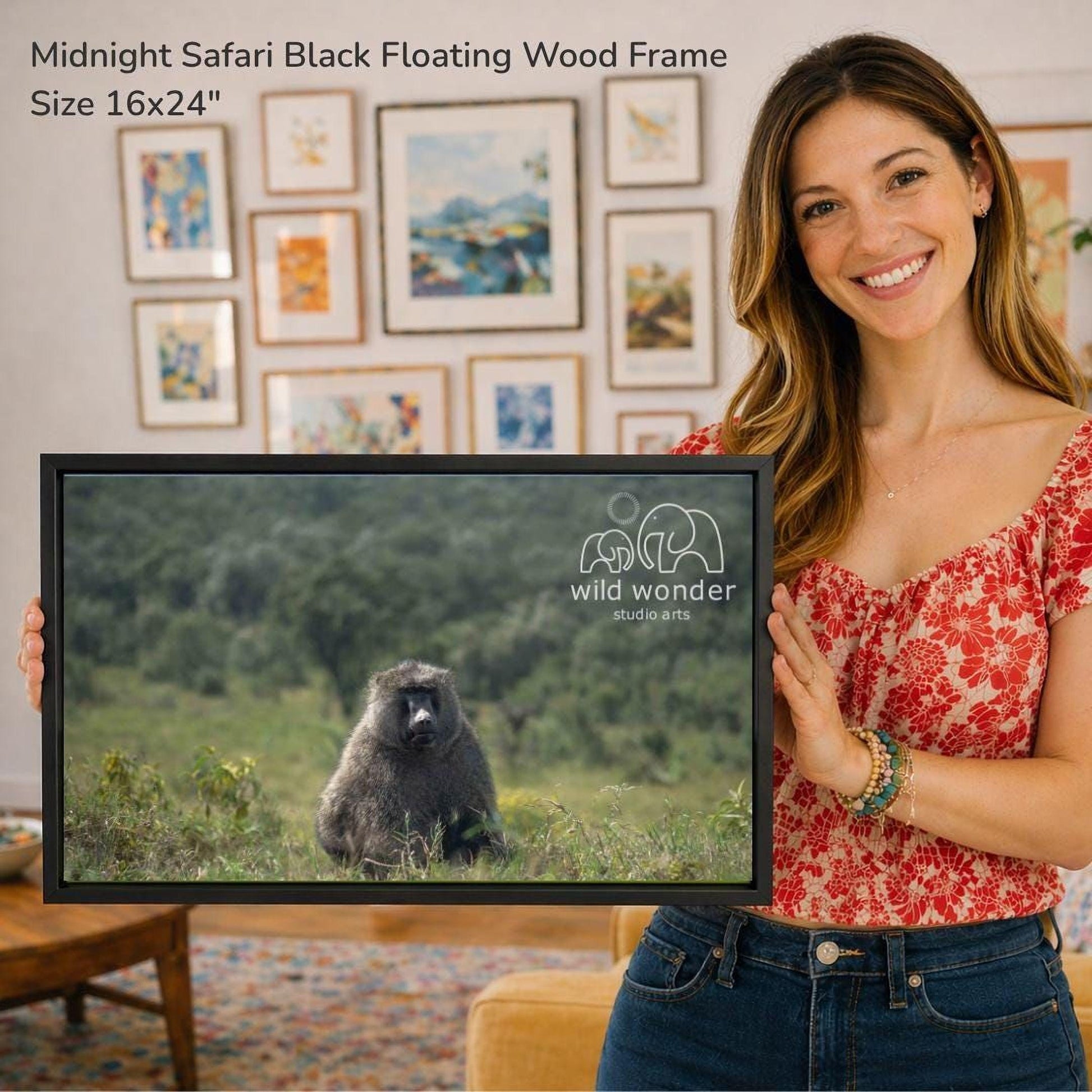 A woman is holding a framed photograph of a black animal, likely a monkey or gorilla, in a grassy field. The framed image is a 16x24" size and is displayed in front of a wall with various framed artworks.