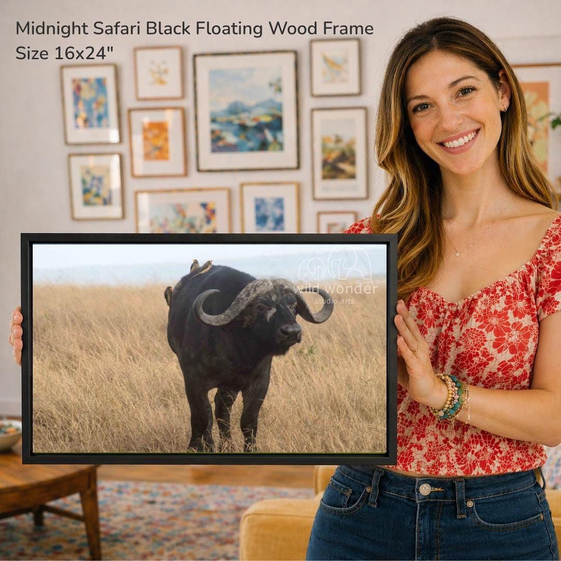 A woman is holding a framed photograph of a black buffalo in a grassy field, with a wall of framed artwork in the background.