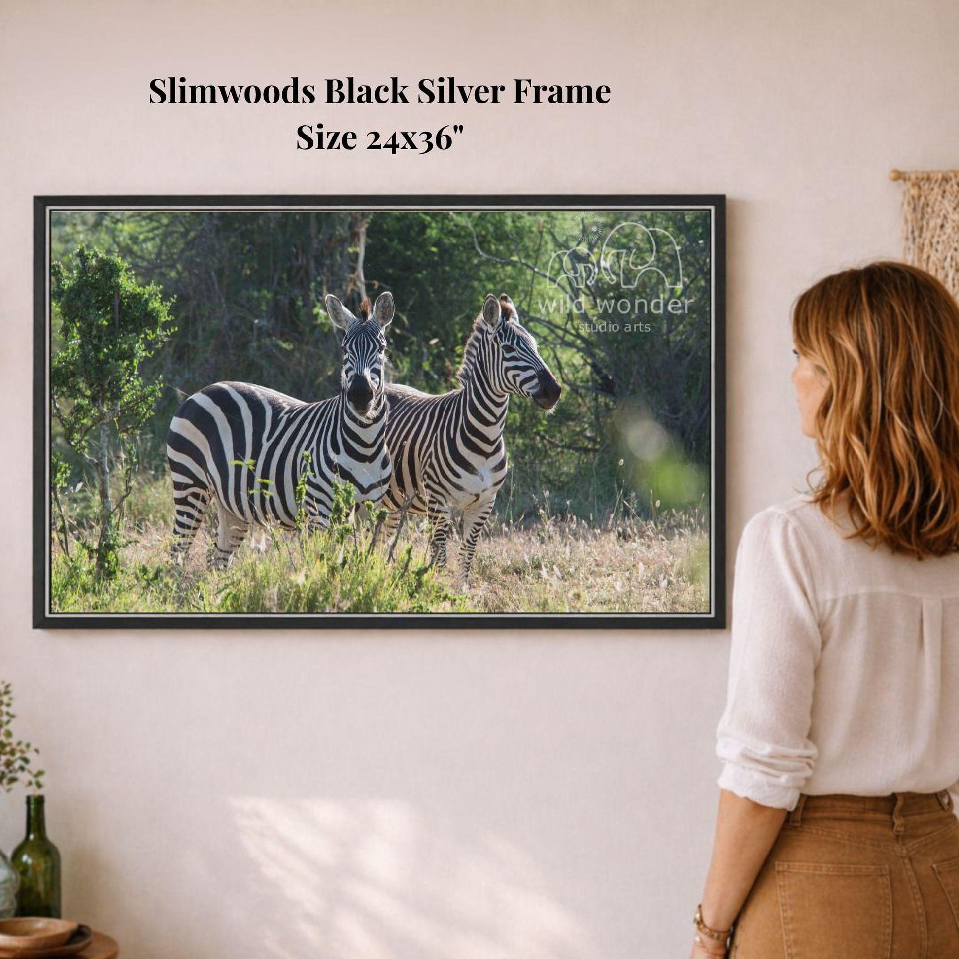 A woman is standing in front of a framed photograph of two zebras in a natural setting, looking at the artwork on the wall.