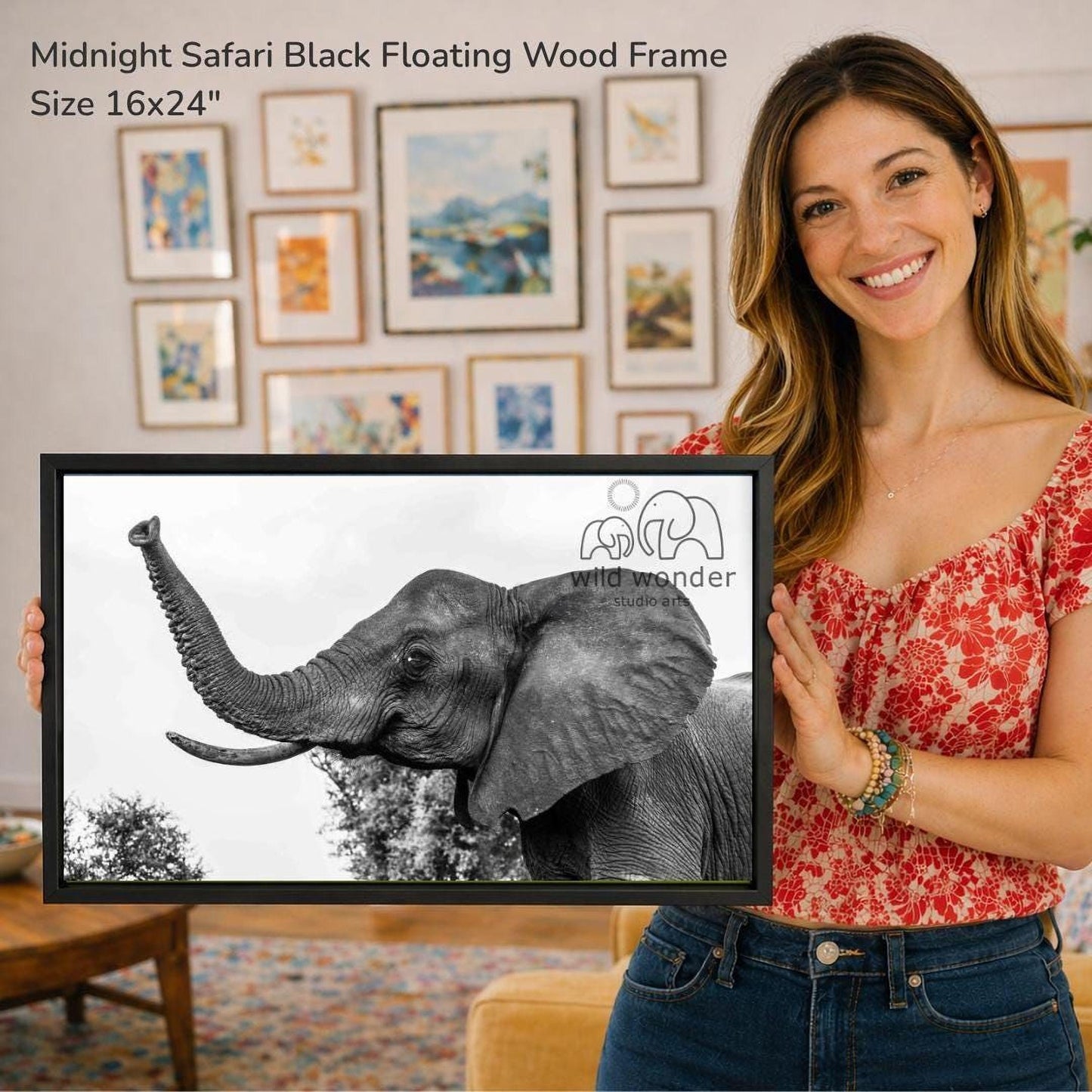 A woman is holding a framed photograph of an elephant in front of a wall with various framed artworks displayed.