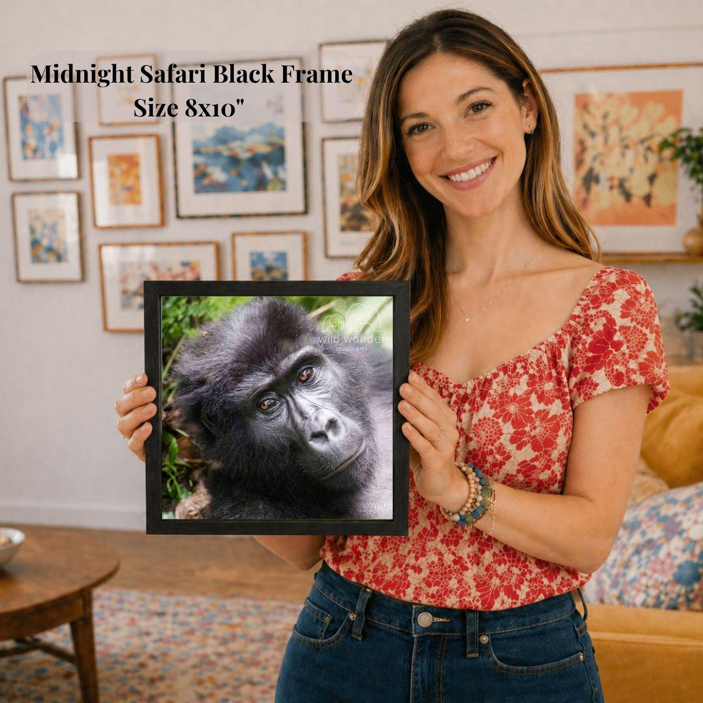 A woman is holding a framed photograph of a gorilla in a forest, smiling and posing for the camera.