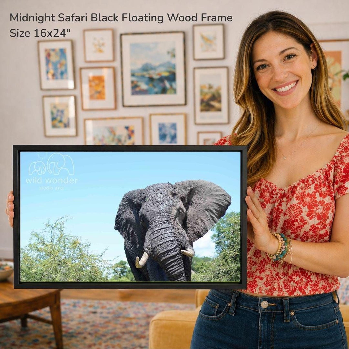 A woman is holding a framed photograph of an elephant in front of a wall with various framed artworks.