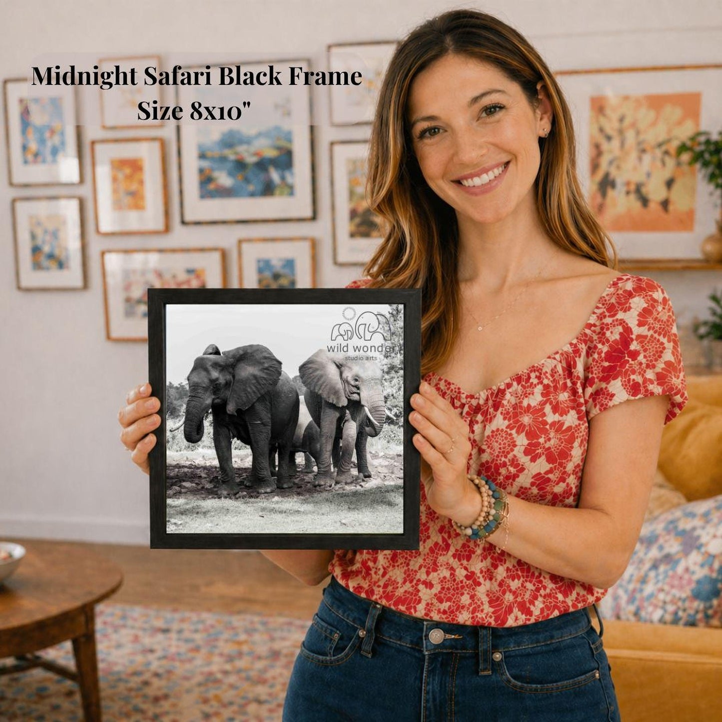 A woman is holding a framed photograph of two elephants in a natural setting, with a smiling expression on her face.