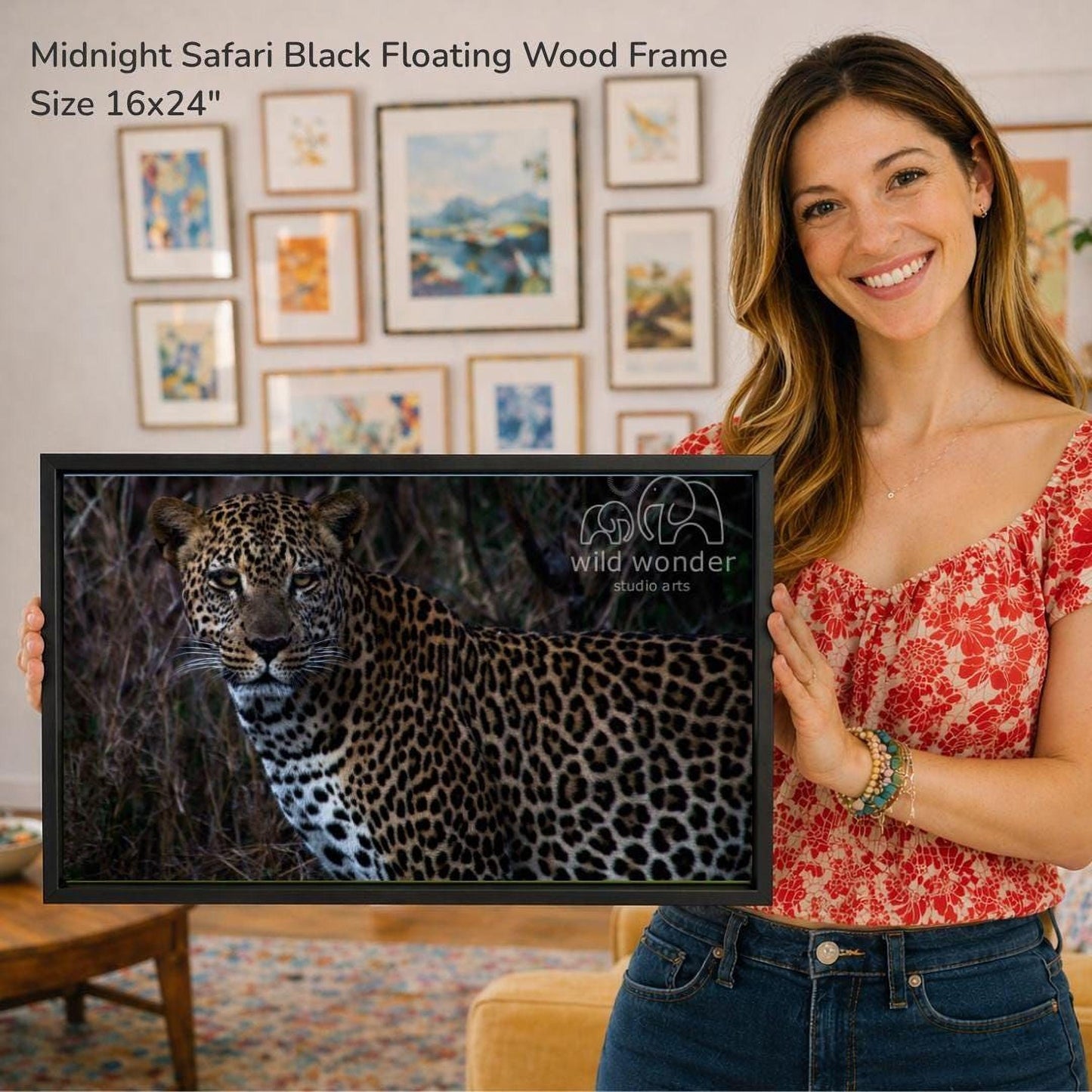 A woman is holding a framed photograph of a leopard in front of a wall with various framed artworks displayed.