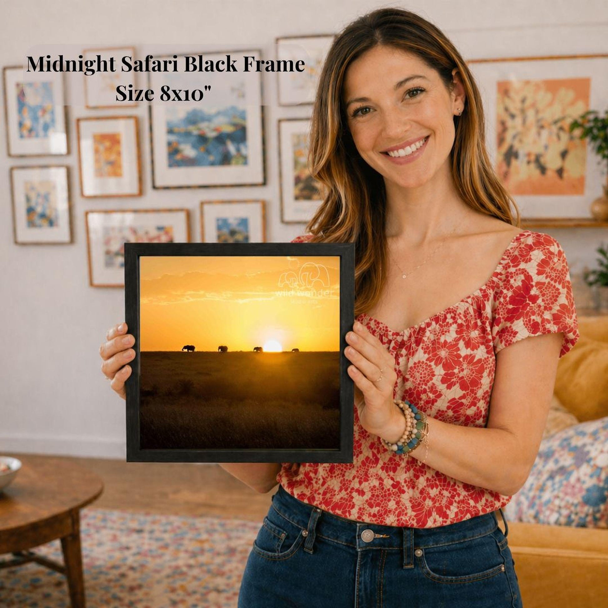 A woman is holding a framed picture of a sunset, smiling and posing in a room with various framed pictures on the wall behind her.