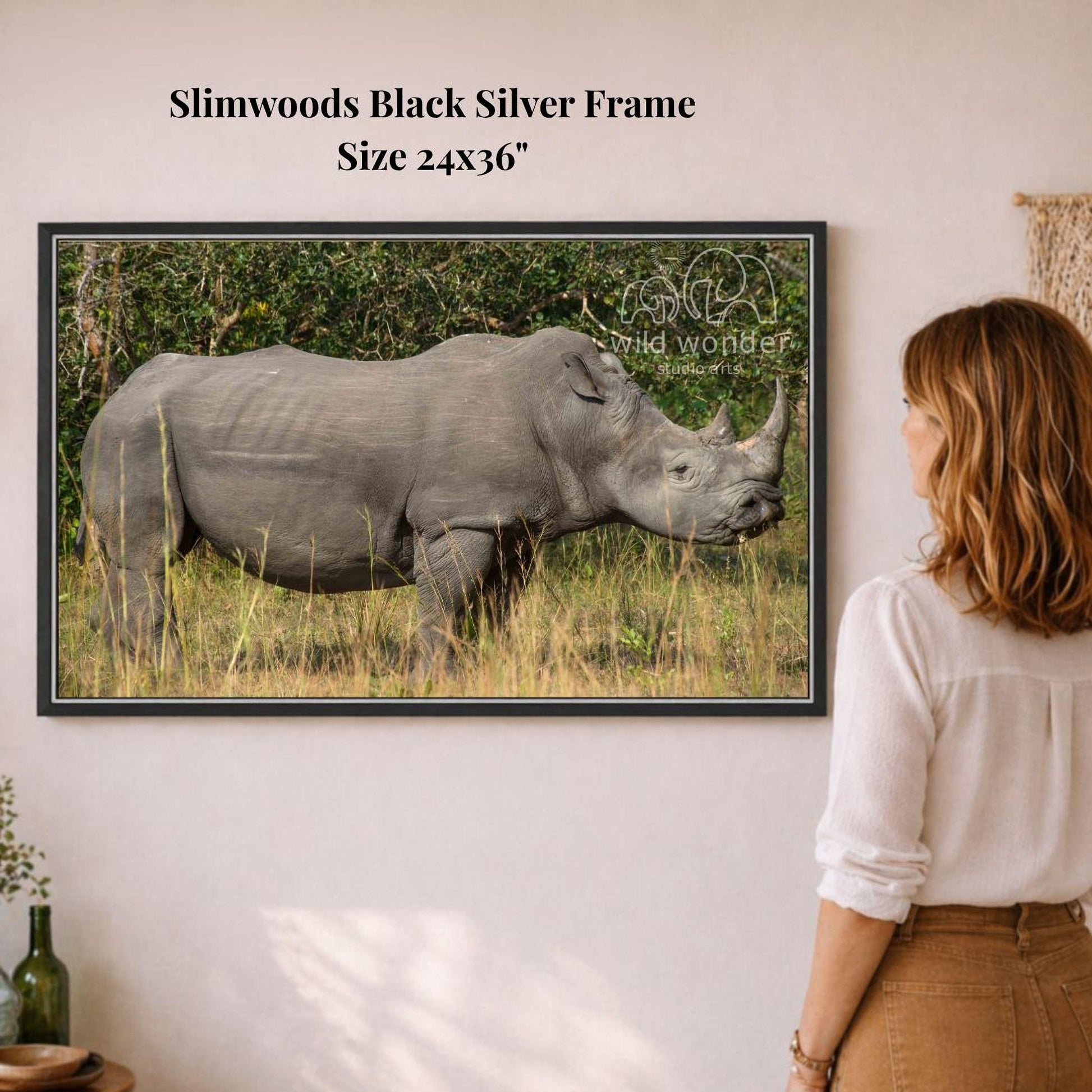 A woman is standing in front of a framed photograph of a rhinoceros in a grassy field, looking at the artwork.
