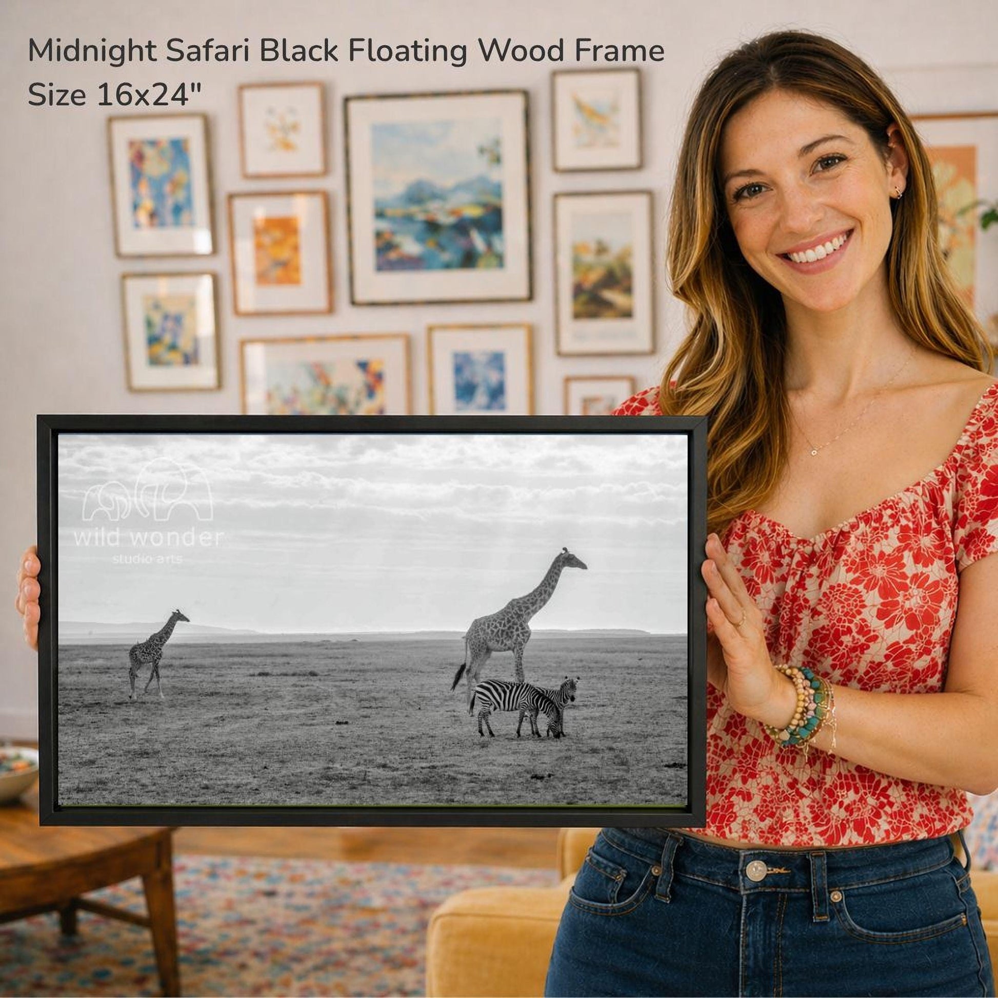 A woman is holding a framed photograph of a giraffe and zebra in a savanna landscape, with a wall of framed art in the background.