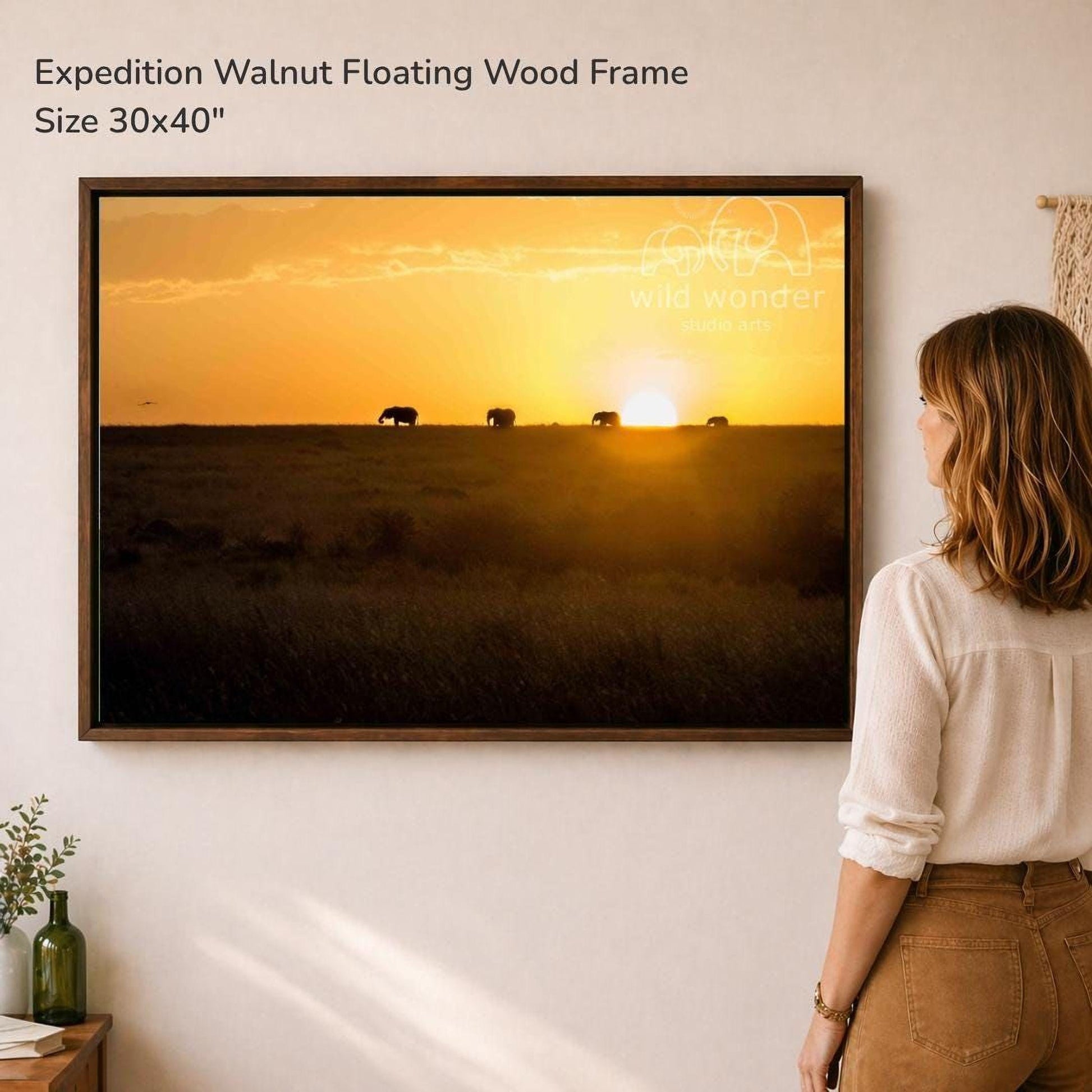 A woman is standing in front of a large framed picture on a wall, looking at it. The picture depicts a sunset over a field with elephants in the distance.