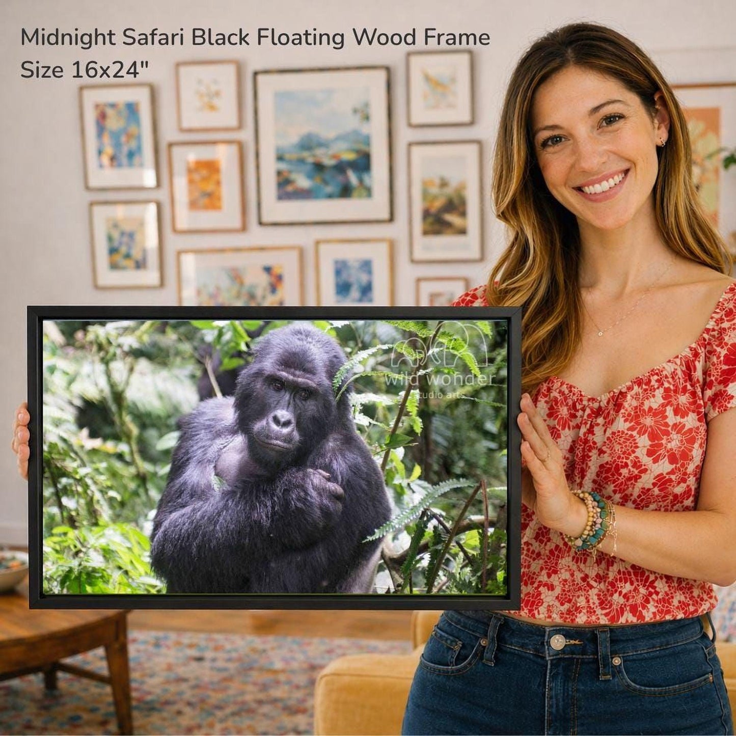 A woman is holding a framed photograph of a gorilla in a forest setting, with a wall of framed art in the background.