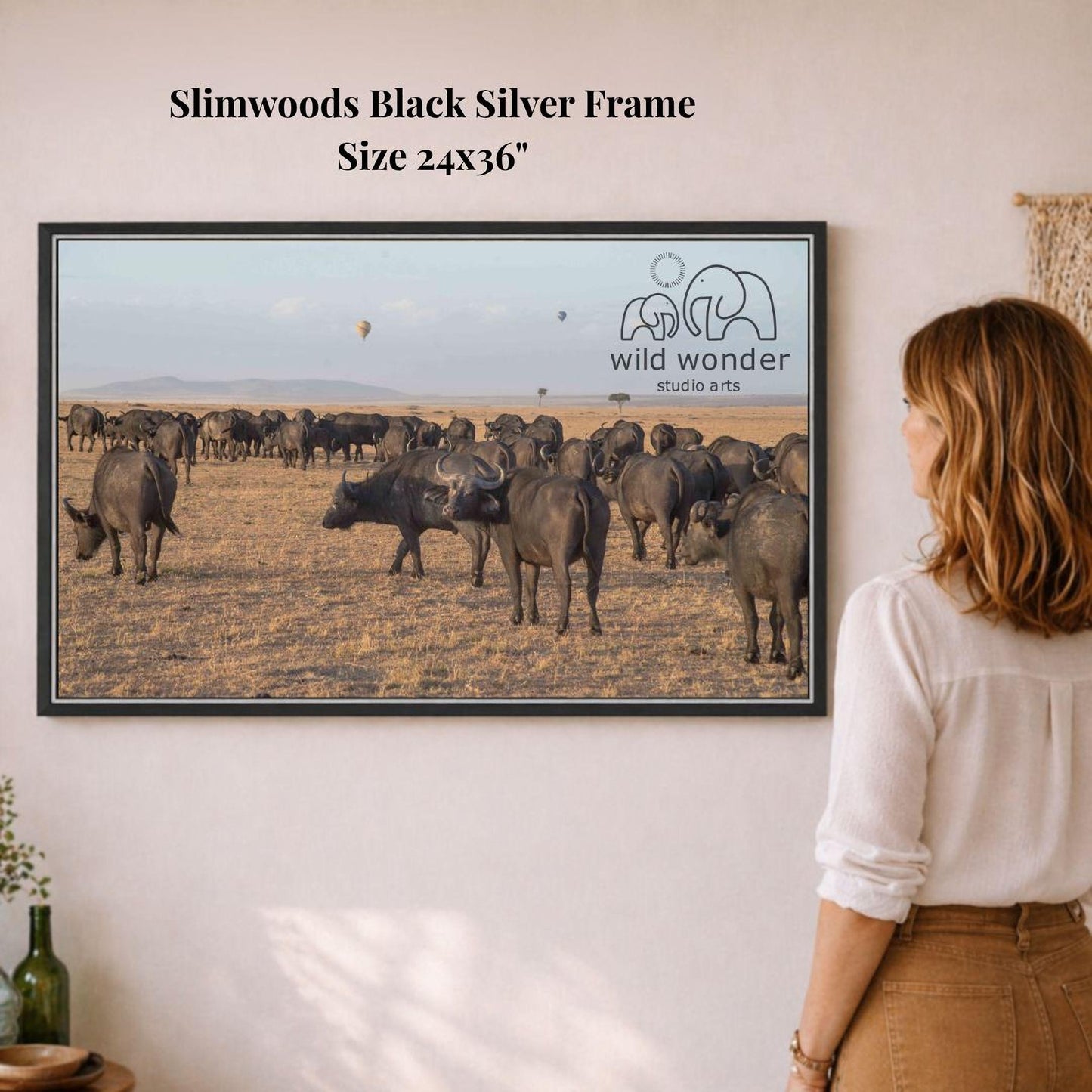 A woman is standing in front of a framed photograph of a herd of elephants in a dry, grassy field.