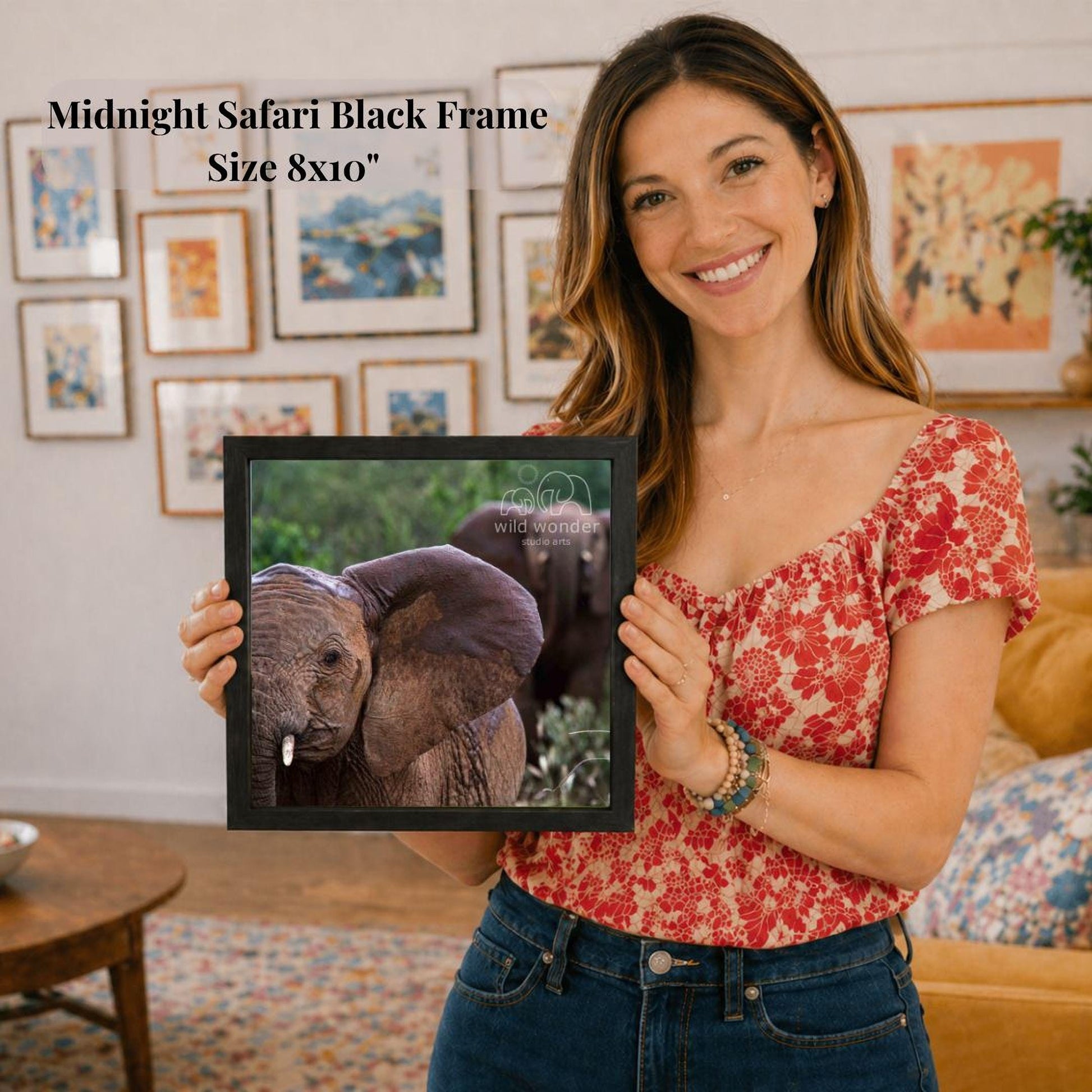 A woman is holding a framed photograph of an elephant in a room with various framed artworks on the walls.