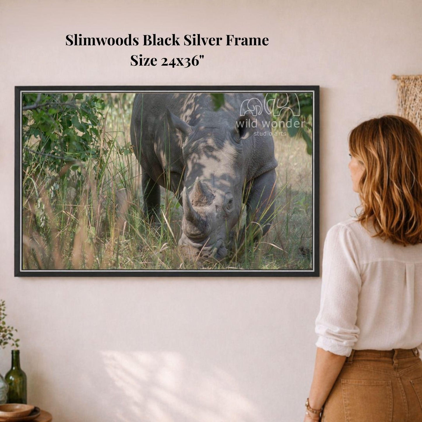 A woman is standing in front of a framed photograph of a rhinoceros in a grassy field, looking at the artwork on the wall.
