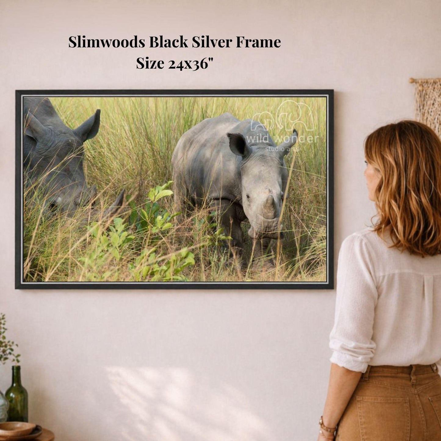 A woman is standing in front of a framed photograph of two rhinos in a grassy field, observing the animals.