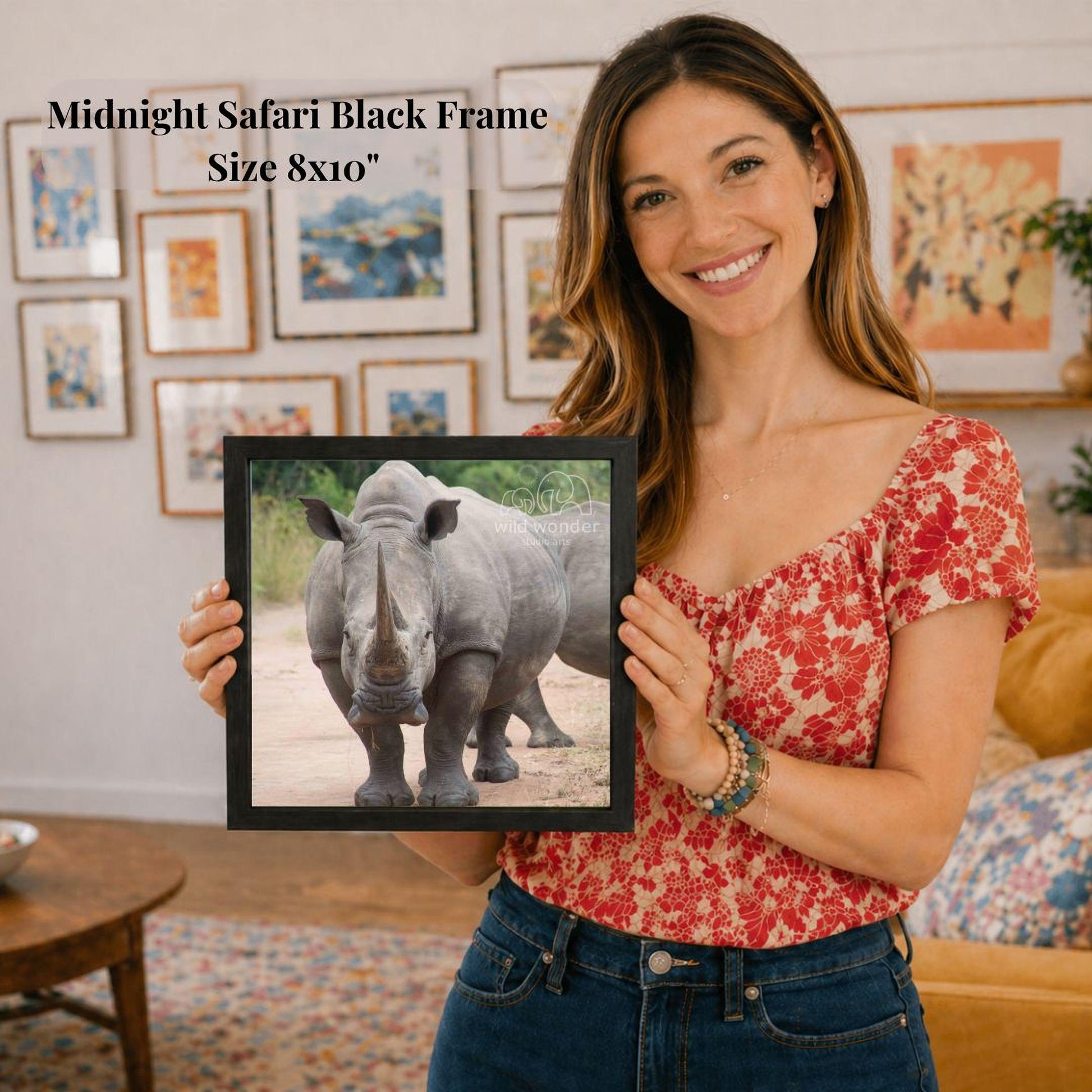 A woman is holding a framed photograph of a rhinoceros in a living room, smiling and posing for the camera.