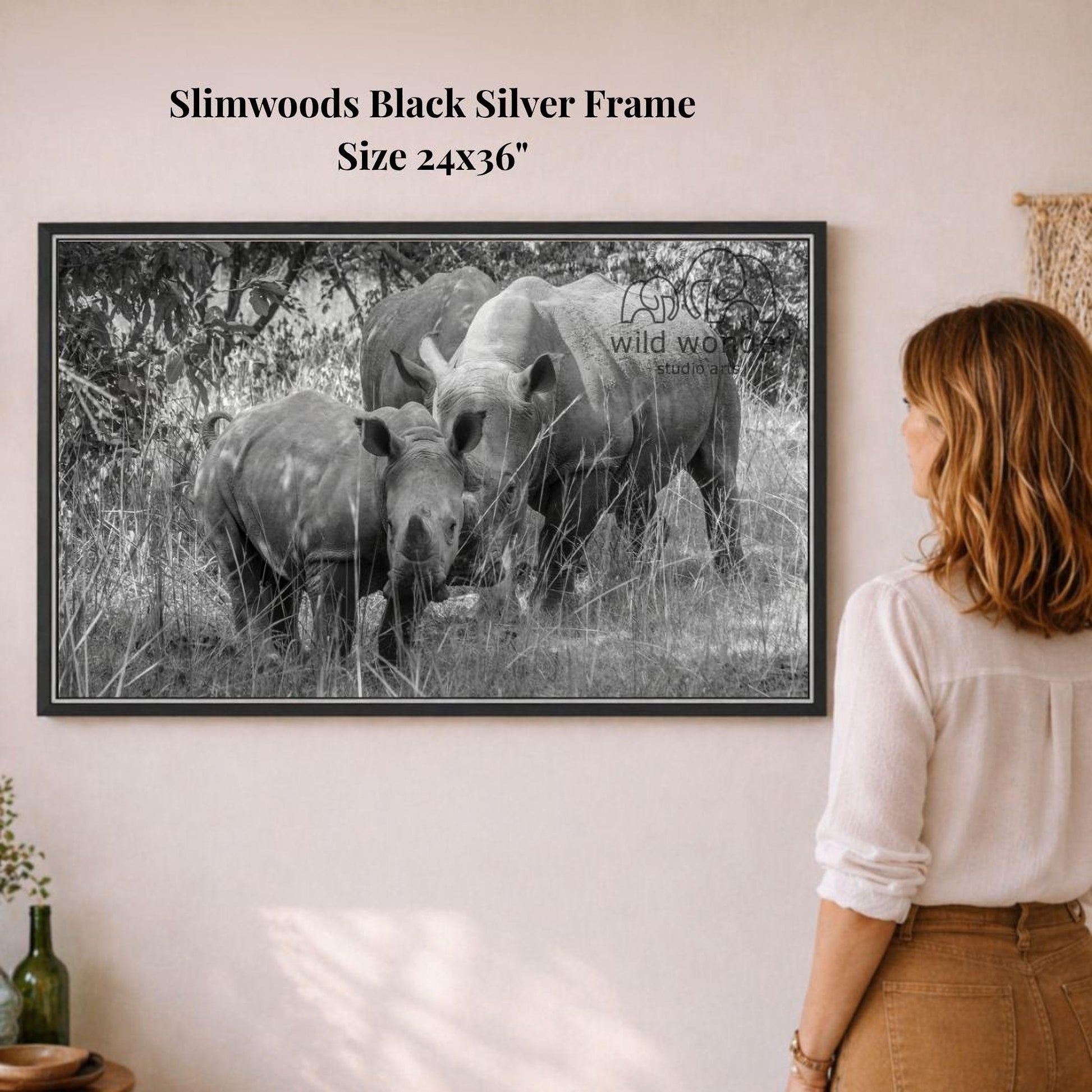 A woman is standing in front of a framed black and white photograph of two rhinos in a grassy field.