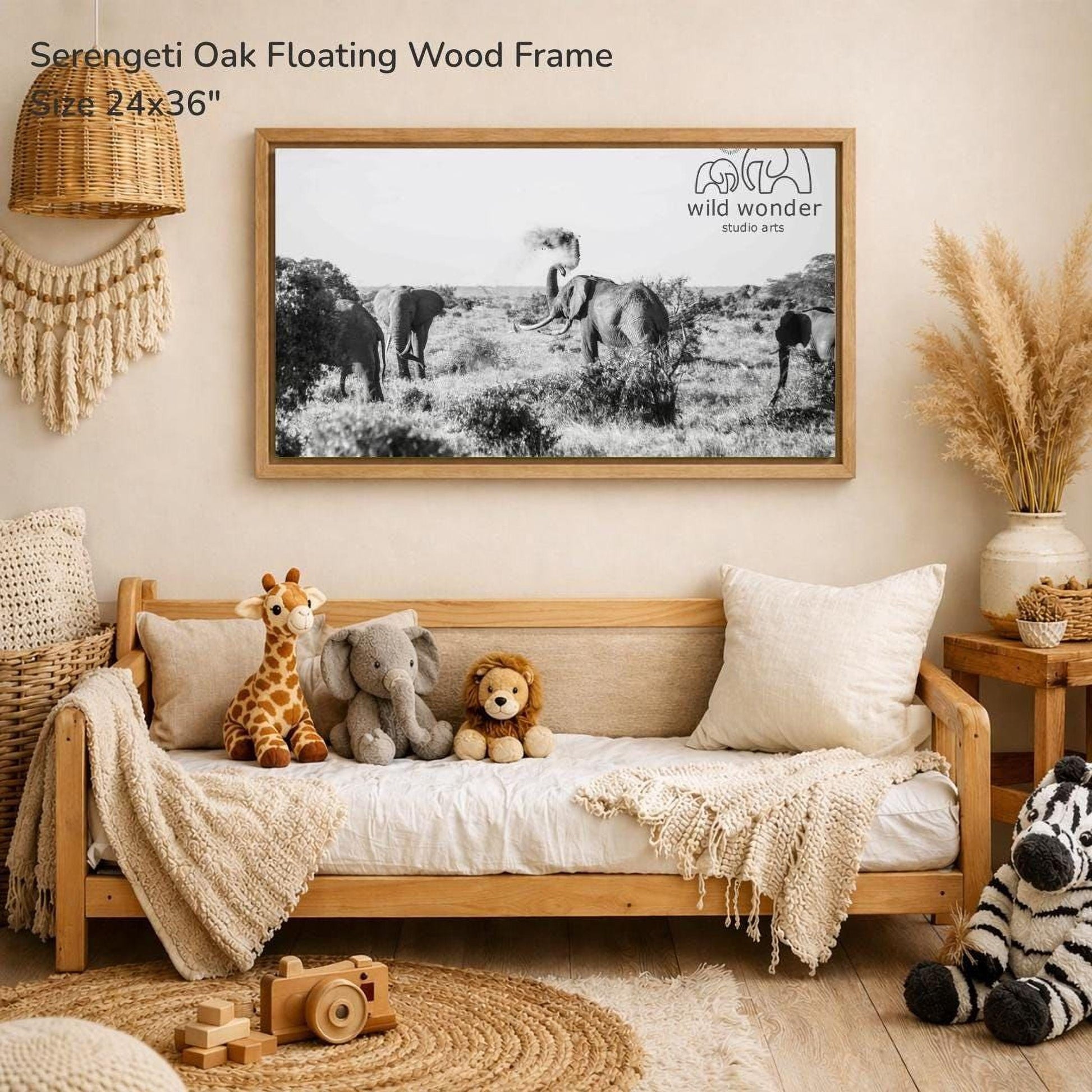 a serene indoor setting with a wooden frame hanging on a wall, displaying a black and white photograph of elephants in a field. The frame is described as a "Serengeti Oak Floating Wood Frame" and measures 24x36 inches. The room is decorated with various items, including a couch with stuffed animals, a wooden toy, and a woven basket.