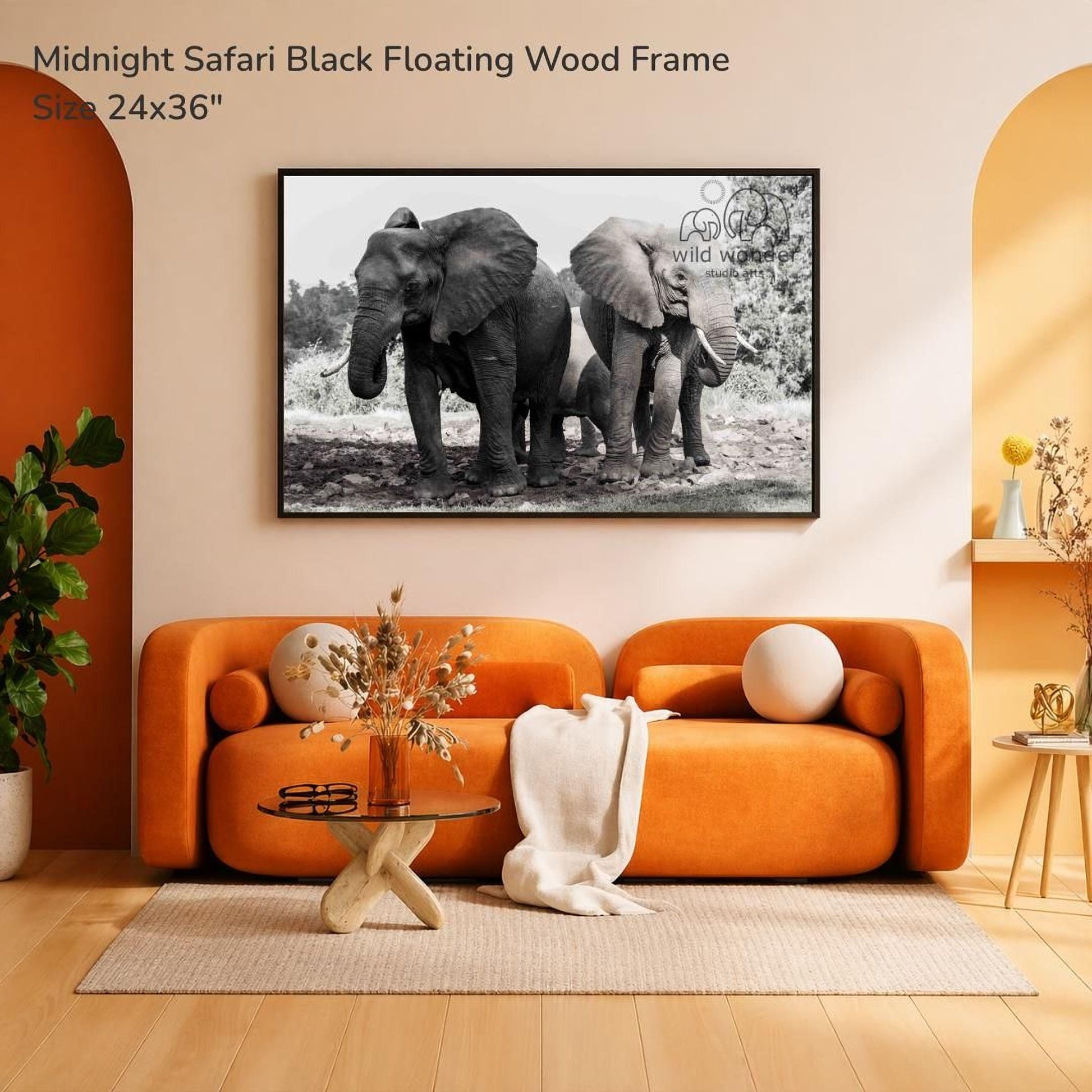 a large black and white photograph of three elephants standing together in a natural setting, mounted on a wall above an orange sofa in a living room.