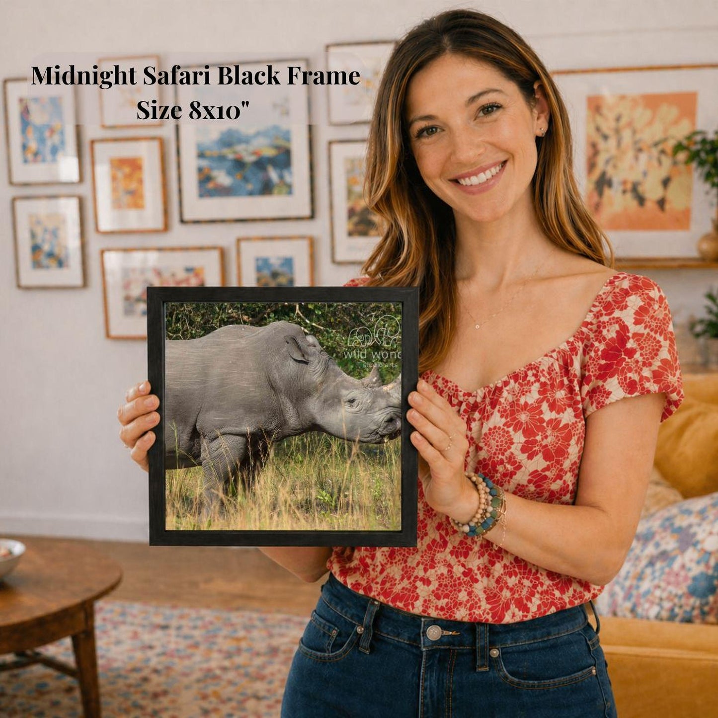 A woman is holding a framed picture of a rhinoceros in a grassy field, smiling and posing for the camera.