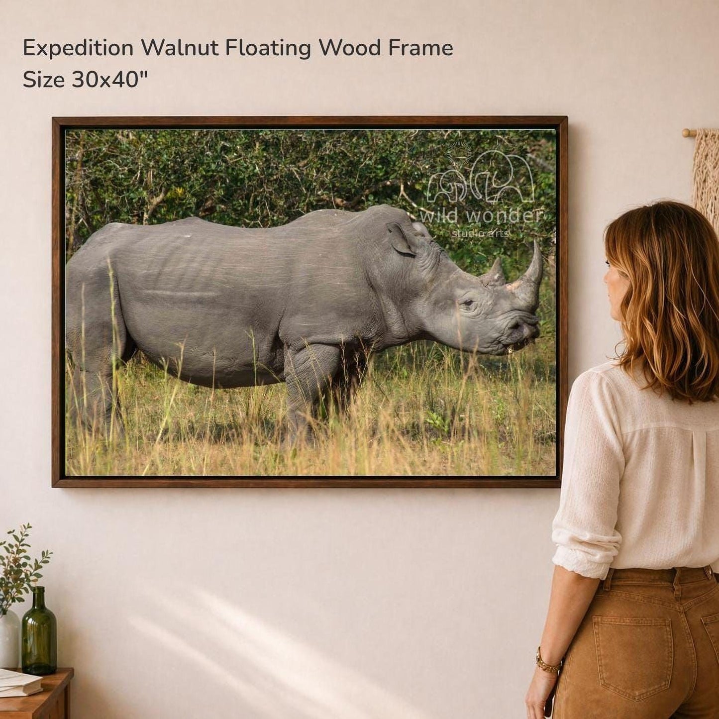 A woman is standing in front of a large framed photograph of a rhinoceros in a grassy field, looking at the artwork.