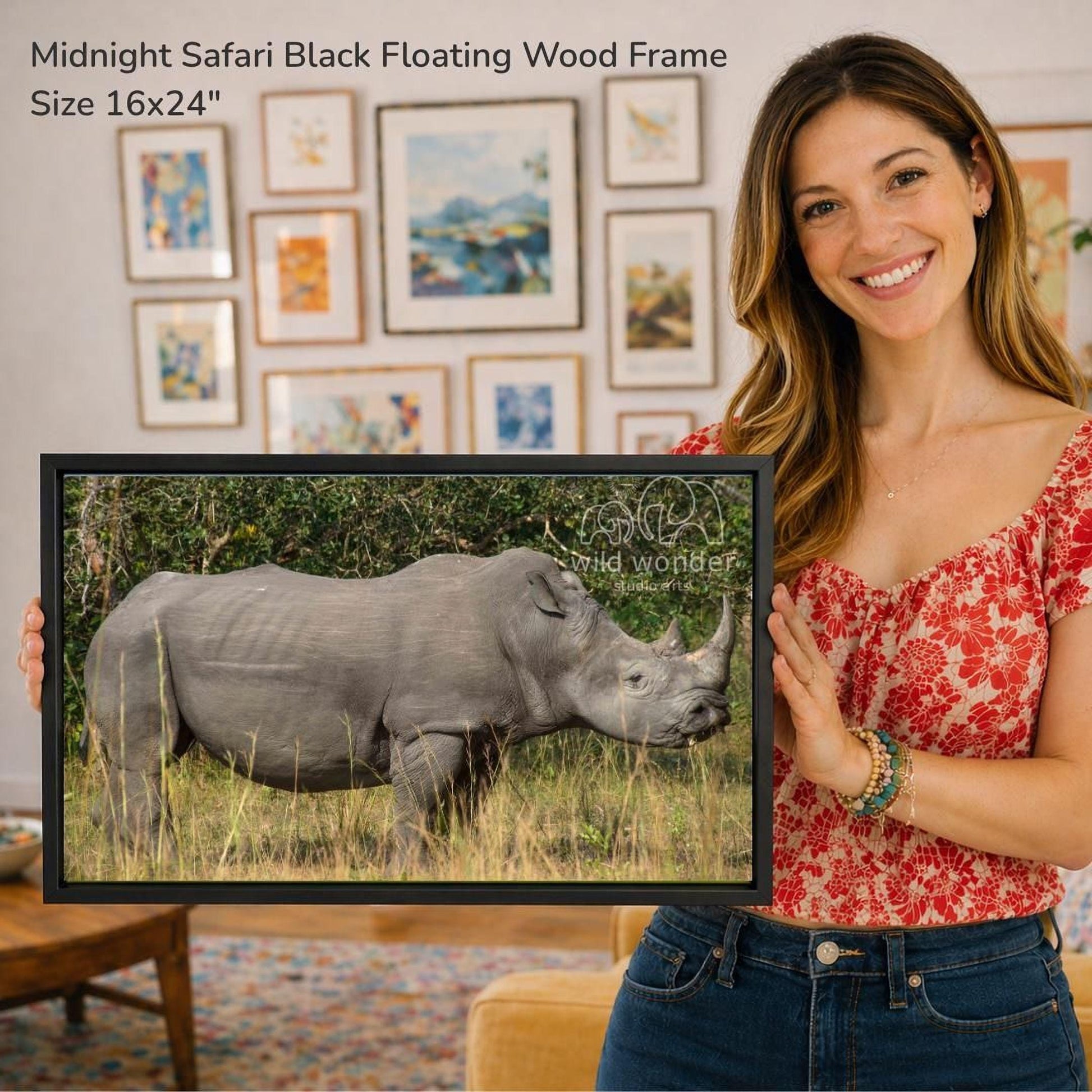 A woman is holding a framed photograph of a rhinoceros in a grassy field, with a smiling expression.