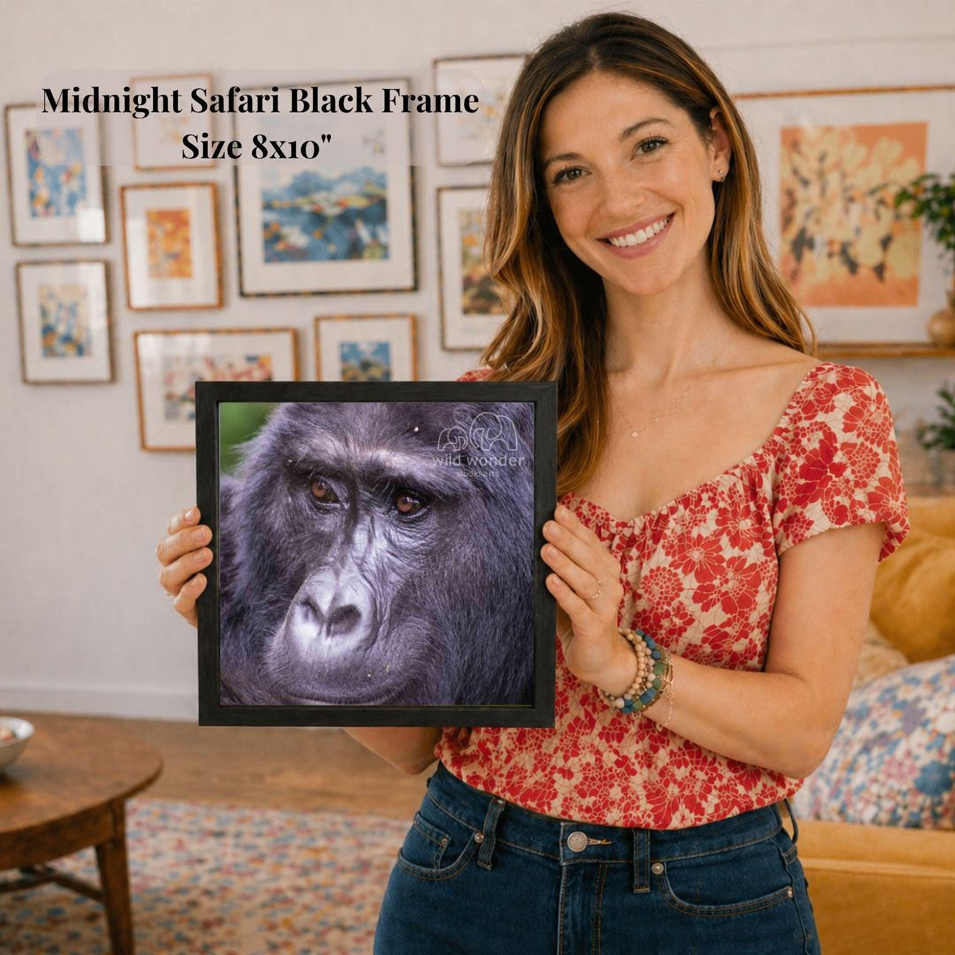 A woman is holding a framed photograph of a gorilla in a room with various framed artworks on the walls.