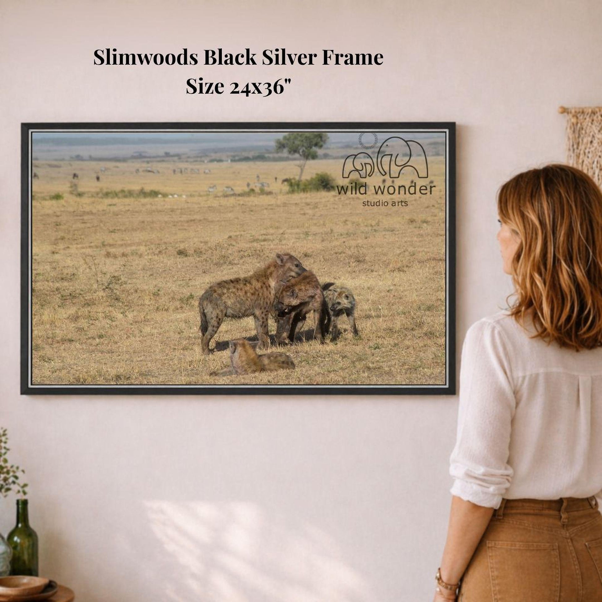 A woman is standing in front of a framed photograph of hyenas in a savanna landscape, looking at the artwork.