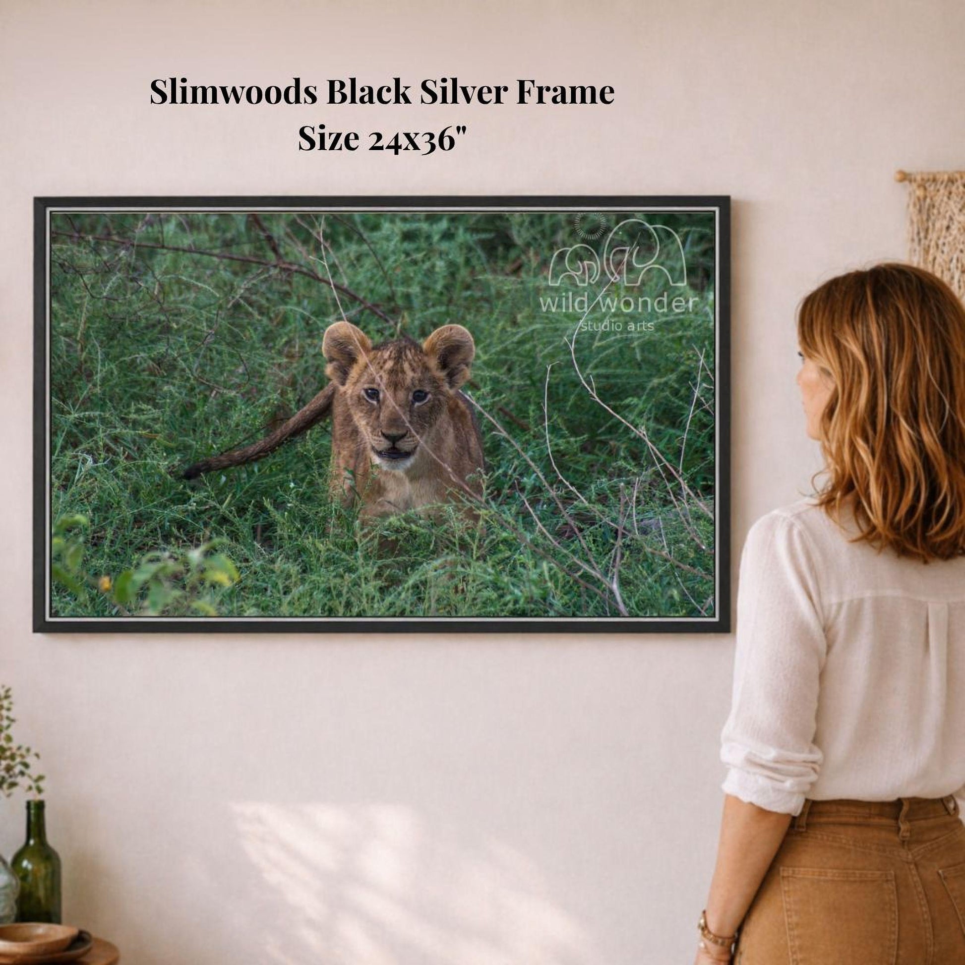 A woman is standing in front of a framed photograph of a young lion cub in a grassy field, looking at the picture.