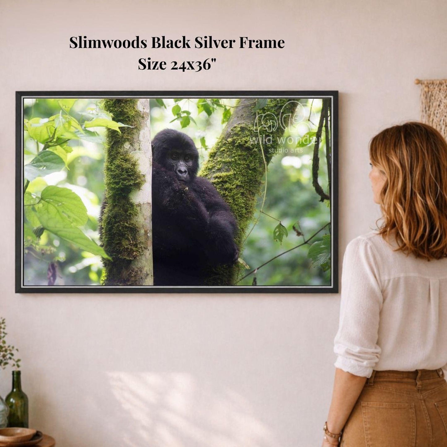 A woman is standing in front of a framed photograph of a gorilla in a lush, green forest, observing the animal.