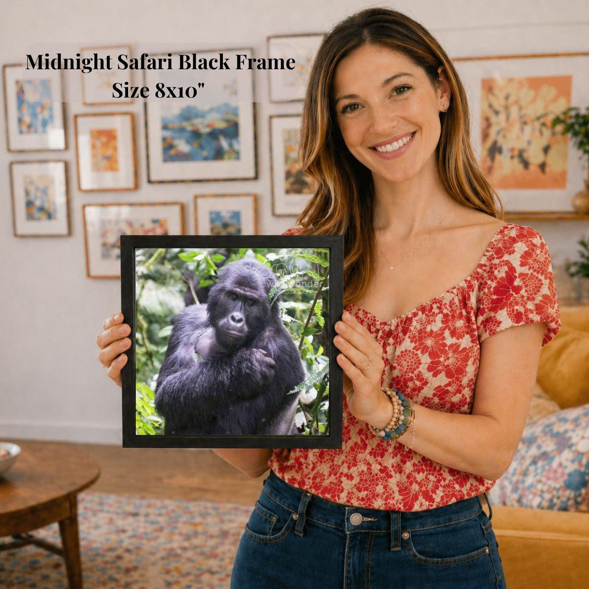 A woman is holding a framed picture of a gorilla in a forest, smiling and posing for the camera.