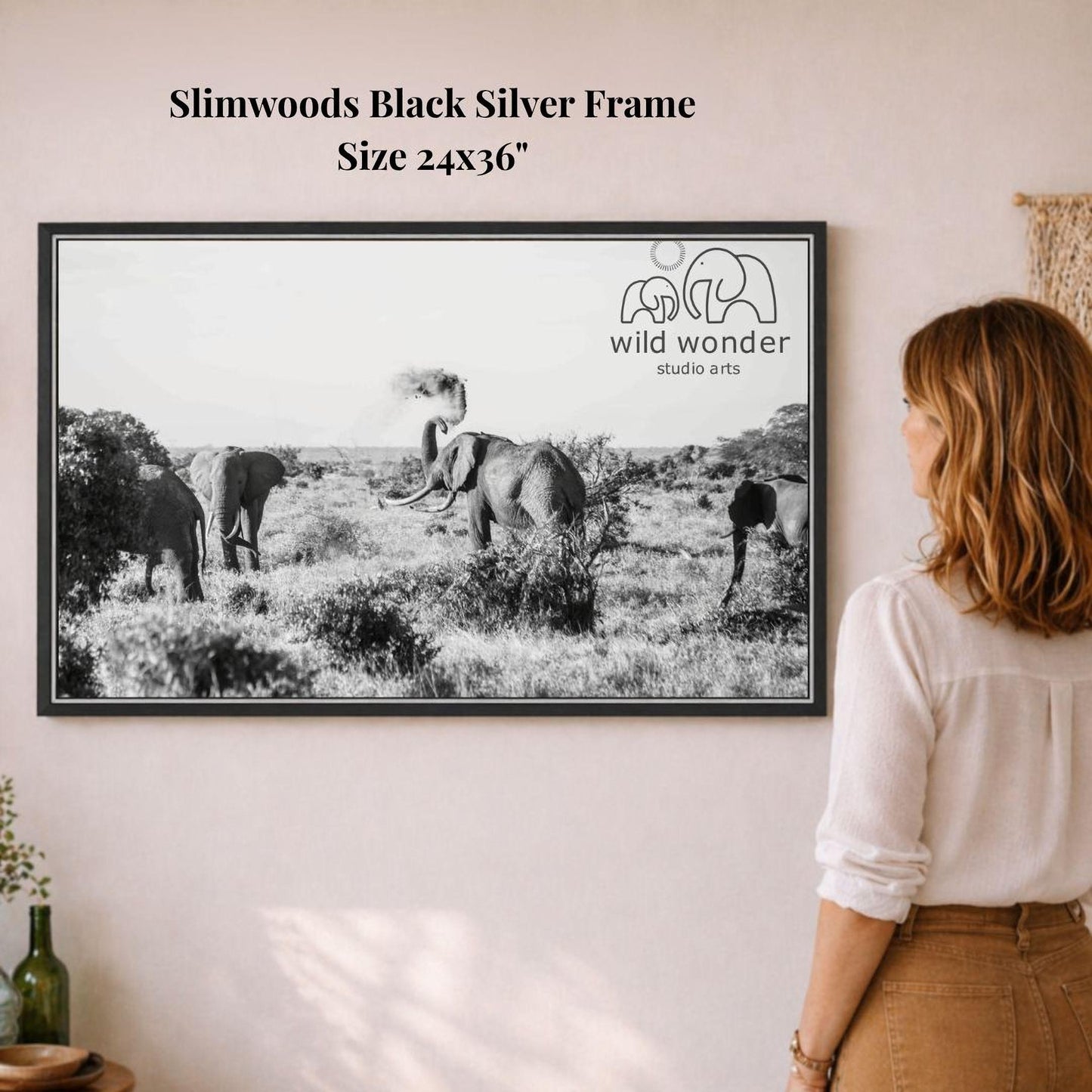 A woman is standing in front of a framed black and white photograph of elephants in a field, looking at the artwork.