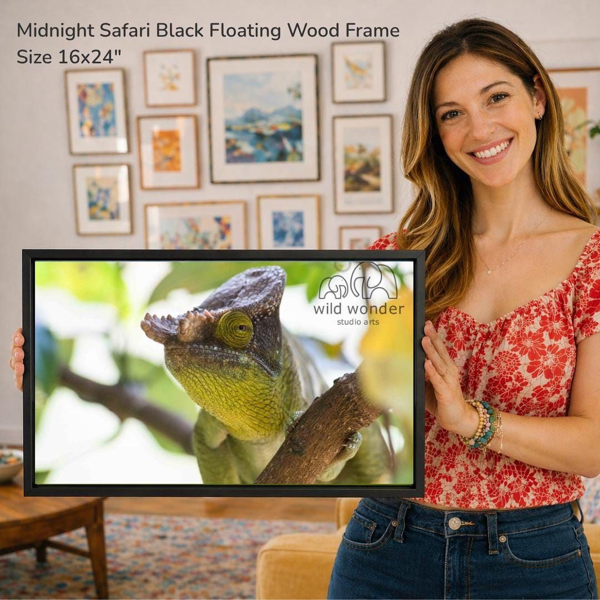 A woman is holding a framed photograph of a chameleon in a wood frame, standing in front of a wall with various framed artworks.
