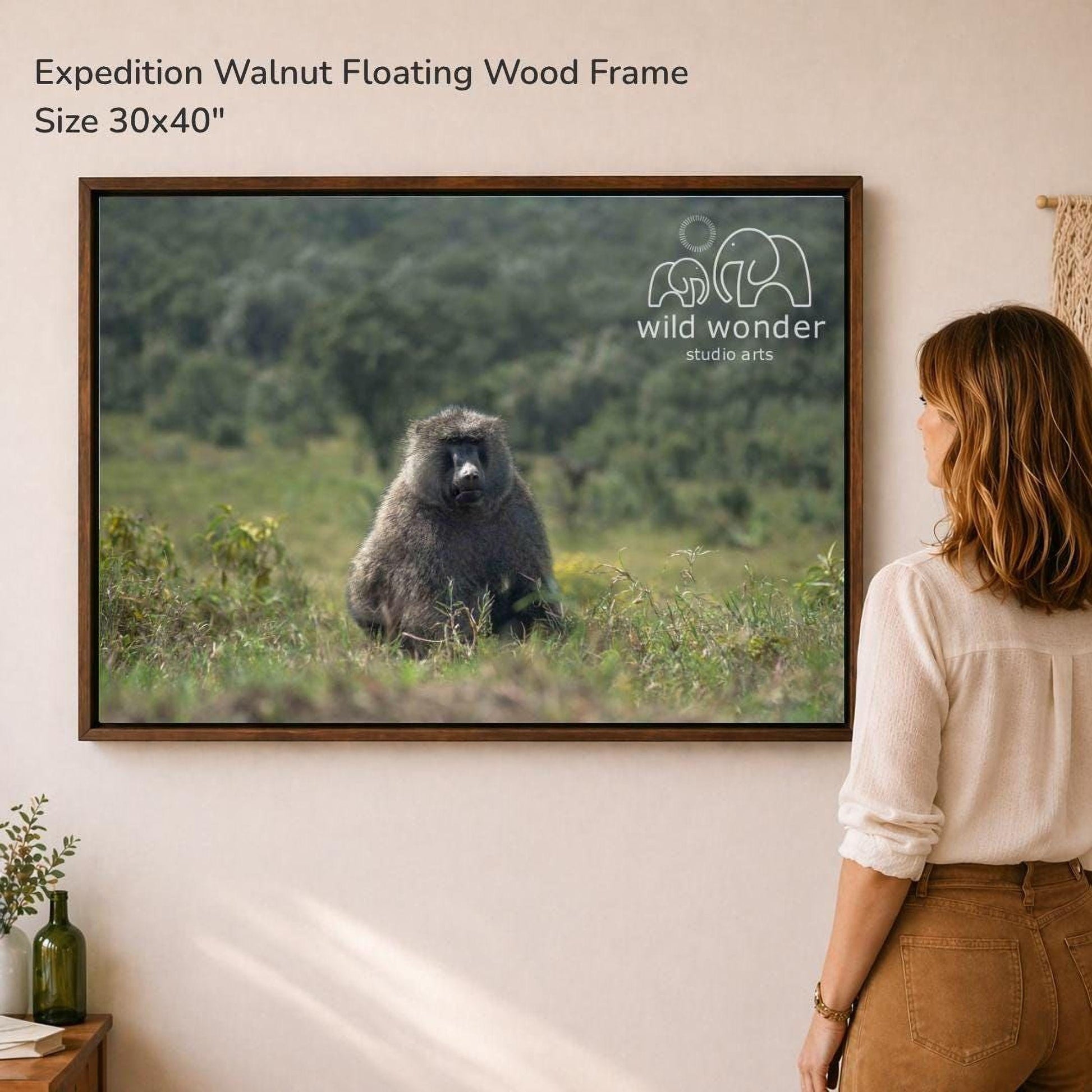 A woman is standing in front of a framed photograph of a baboon in a grassy field, looking at the artwork.