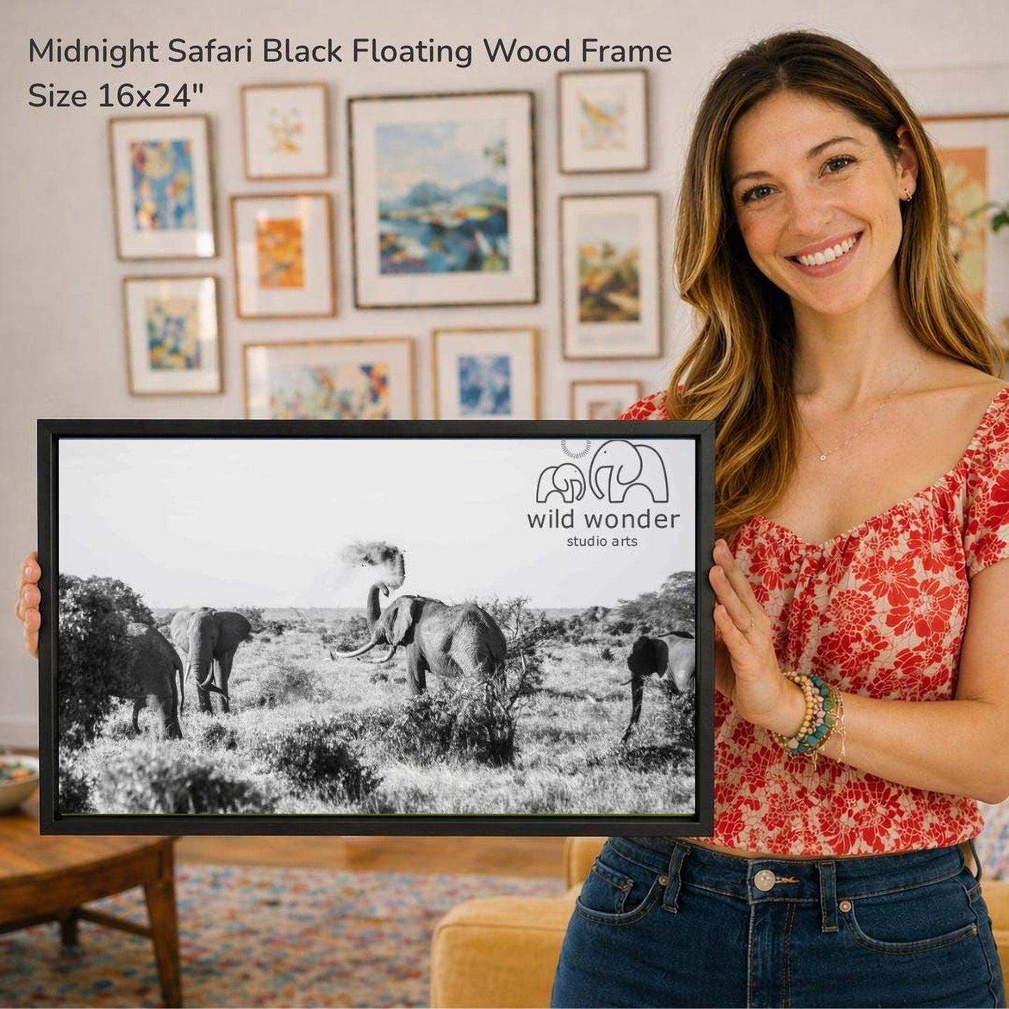 A woman is holding up a framed black and white photograph of elephants in a field, with a smiling expression on her face.