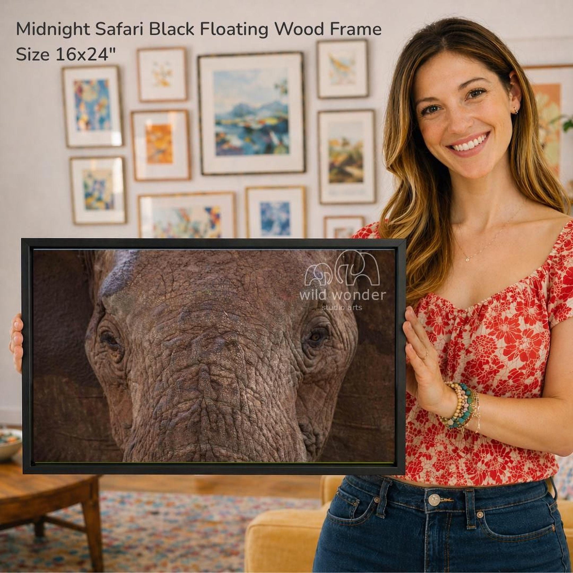 A woman is holding a framed photograph of an elephant in front of a wall with various framed artworks displayed.