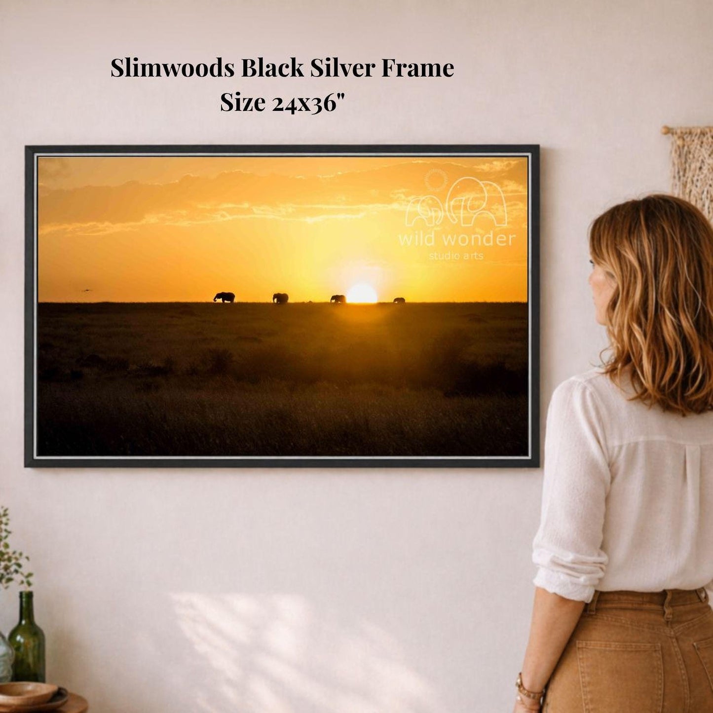A woman is standing in front of a framed photograph of elephants in a field at sunset, looking at the artwork.