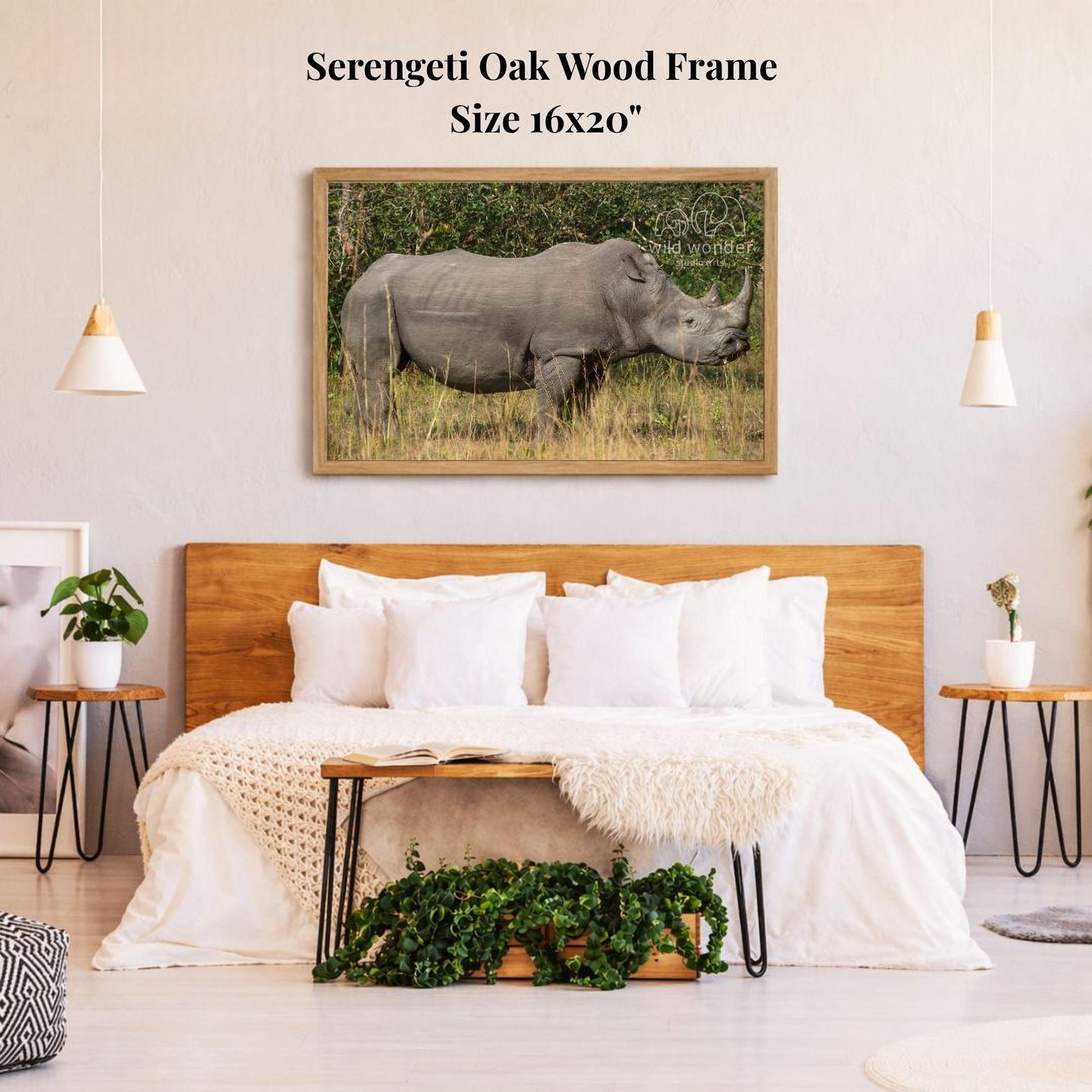 a bedroom with a large wooden headboard and a white bedding set. Above the bed, there is a framed photograph of a rhinoceros in a grassy field. The wall behind the bed has a text that reads "Serengeti Oak Wood Frame Size 16x20".