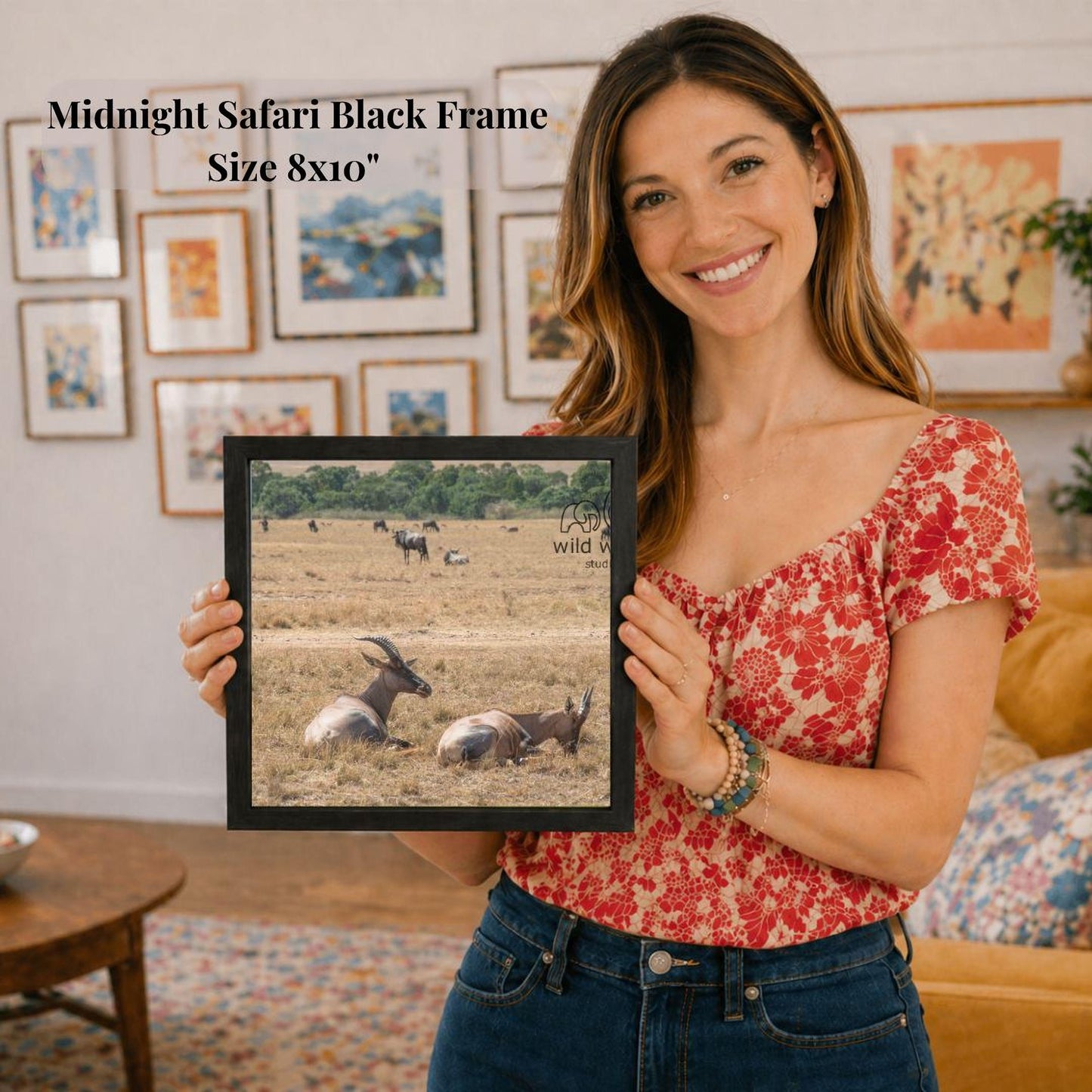 A woman is holding a framed picture of a group of wild animals, including a giraffe and a zebra, in a living room setting.