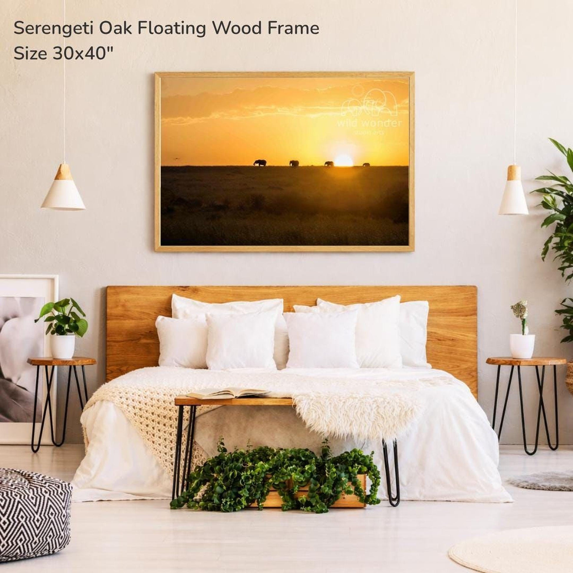 a serene bedroom setting with a large wooden headboard and a cozy bed. Above the bed, there is a framed picture of a sunset with a herd of elephants in the distance. The room is decorated with potted plants and other natural elements, creating a warm and inviting atmosphere.