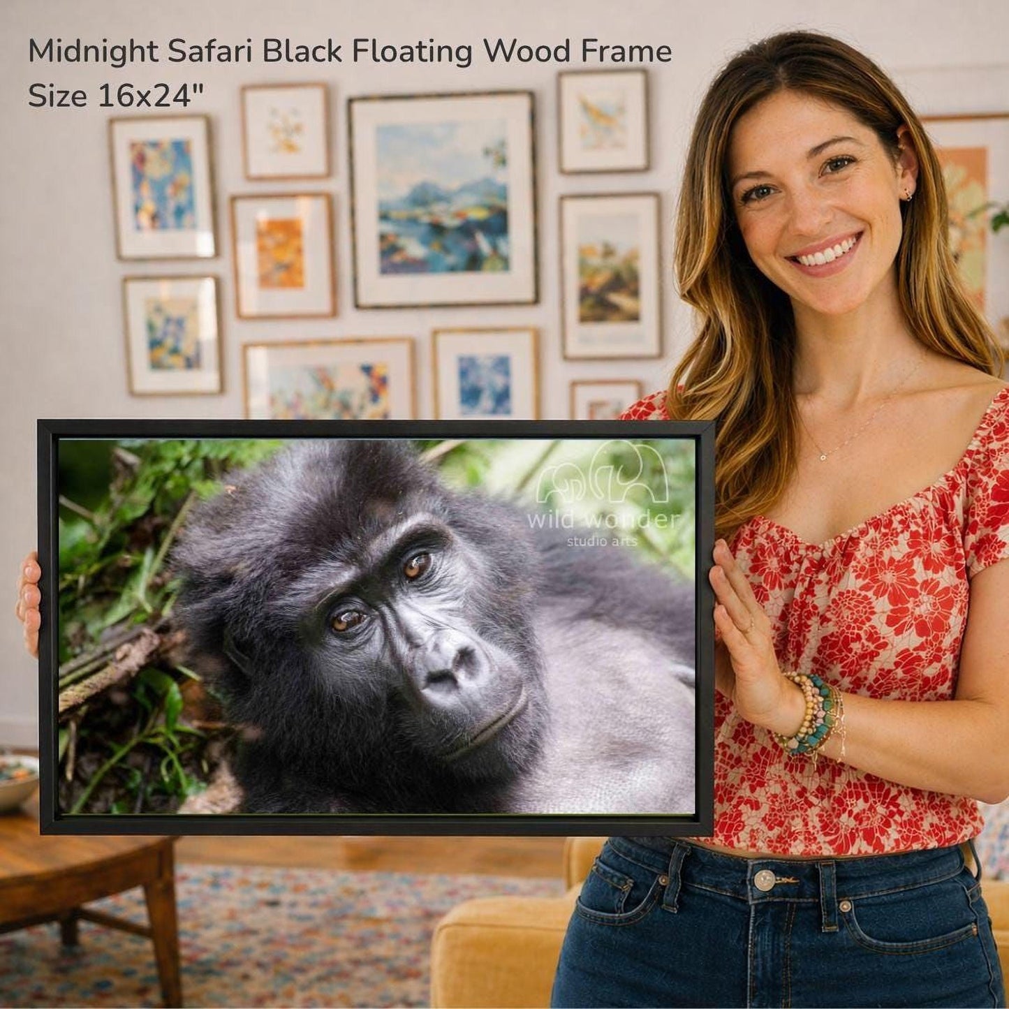 A woman is holding a framed photograph of a gorilla in a forest setting, with a wall of framed art in the background.