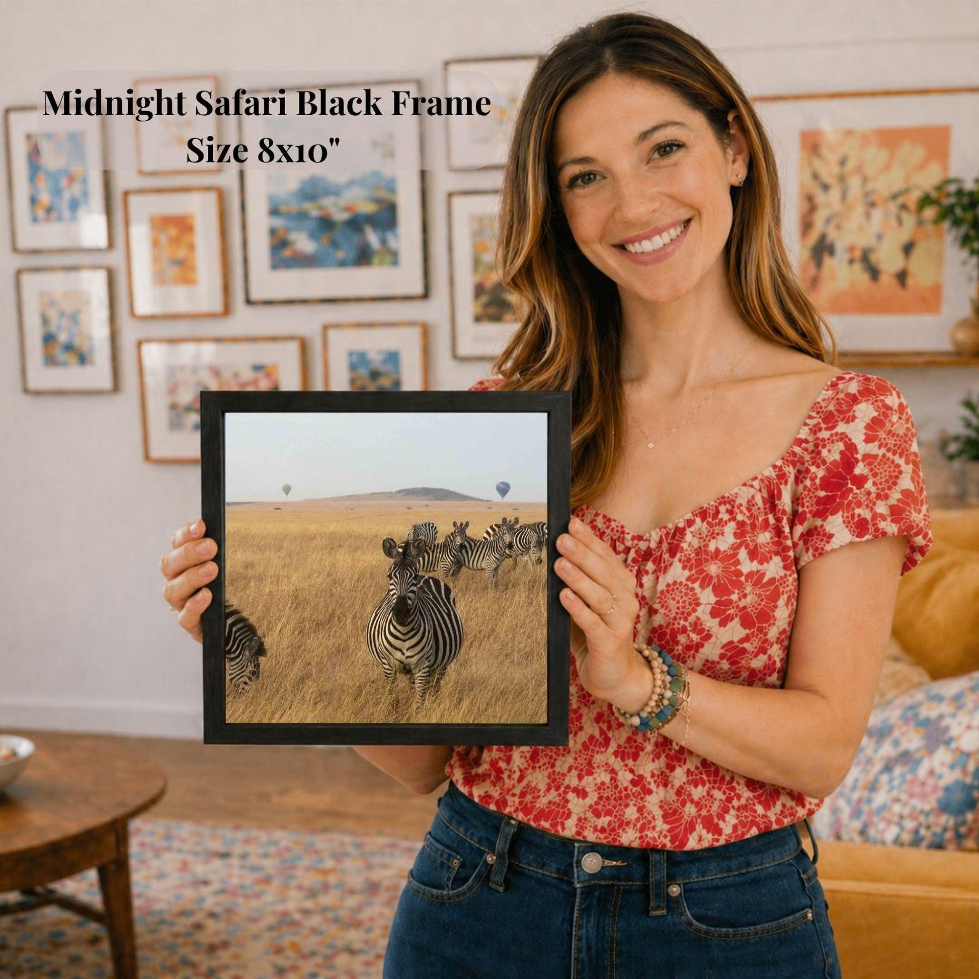 A woman is holding a framed photograph of a group of zebras in a grassy field, with a smiling expression on her face.
