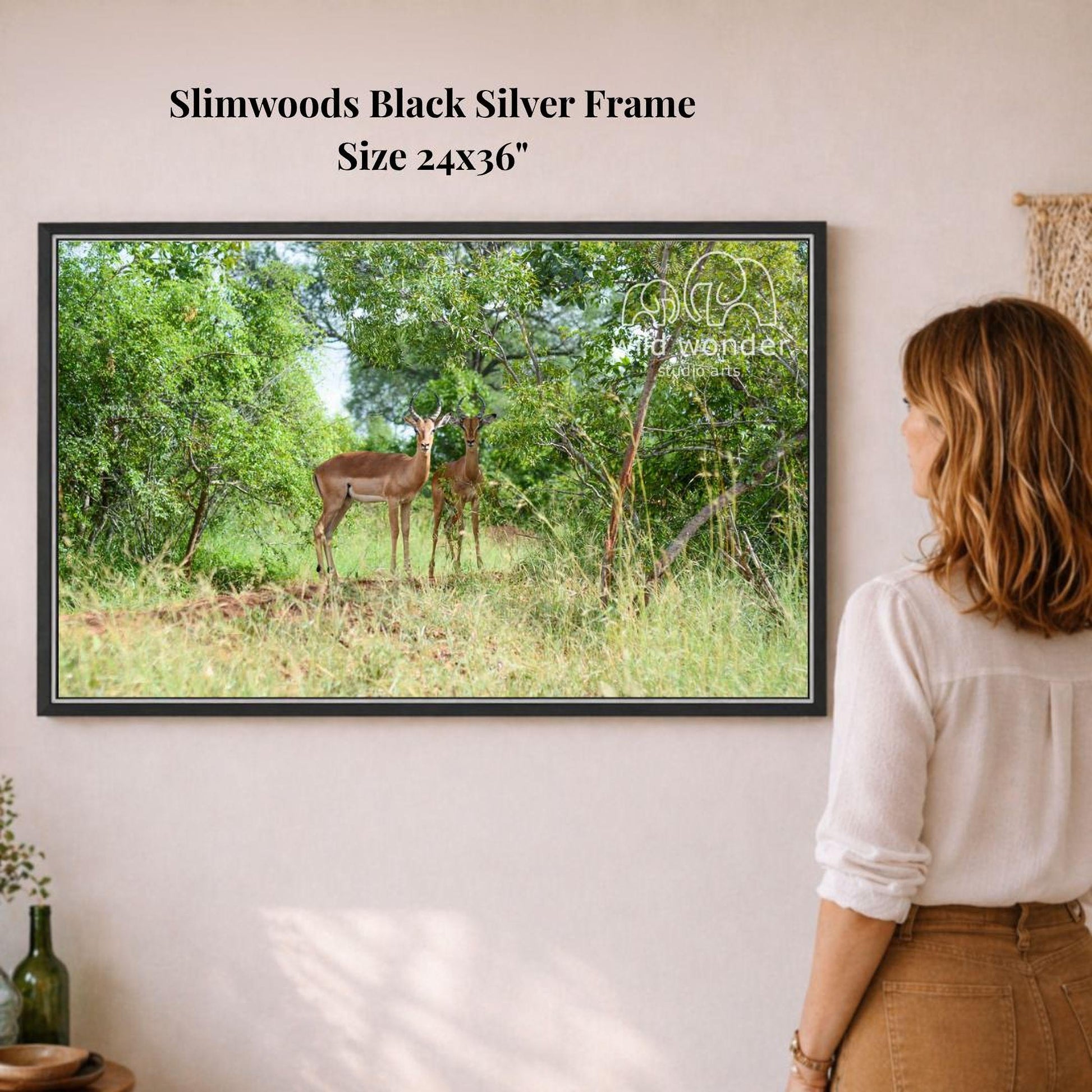 A woman is standing in front of a framed photograph of two deer in a forest, looking at the artwork on the wall.