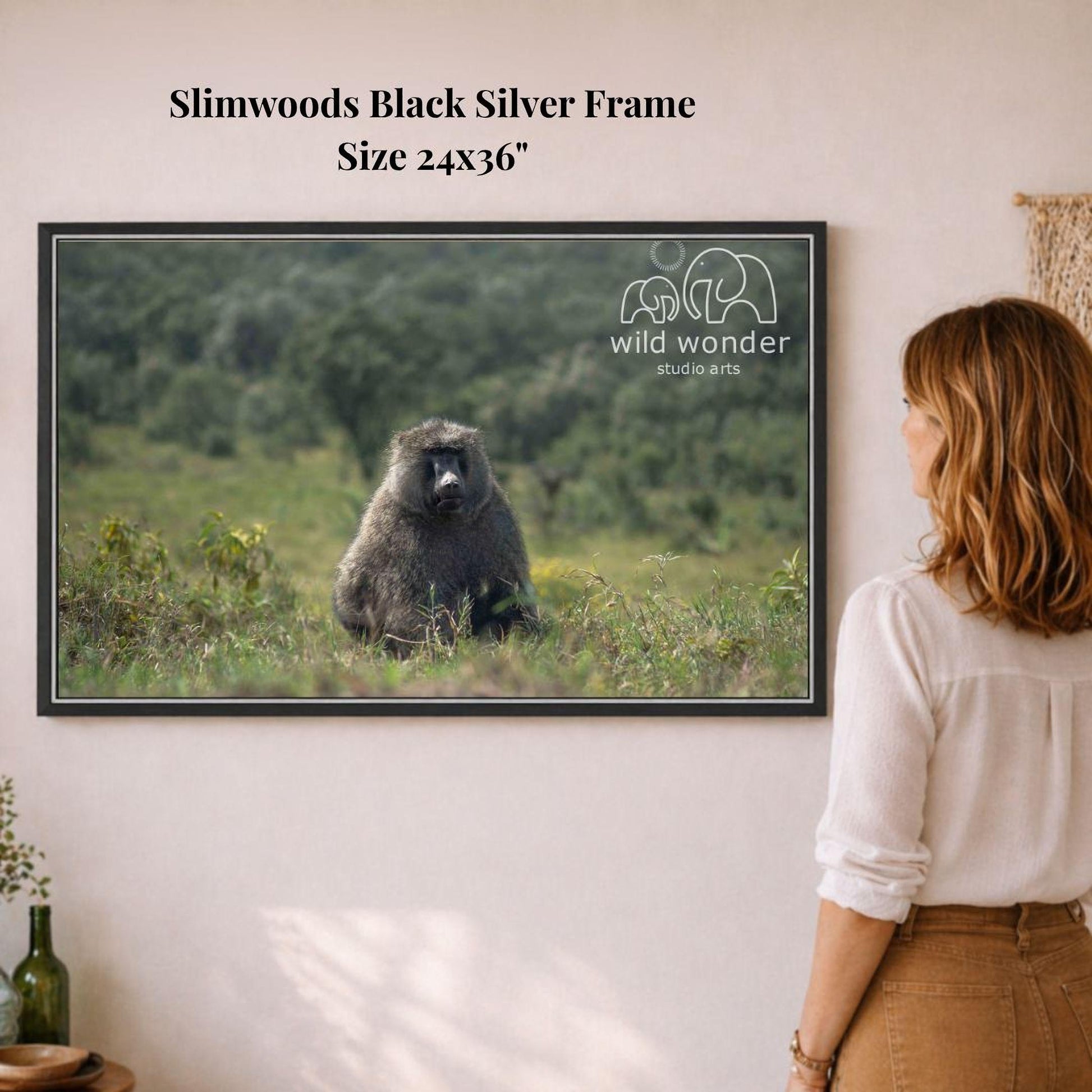 A woman is standing in front of a framed photograph of a baboon in a grassy field, looking at the artwork.
