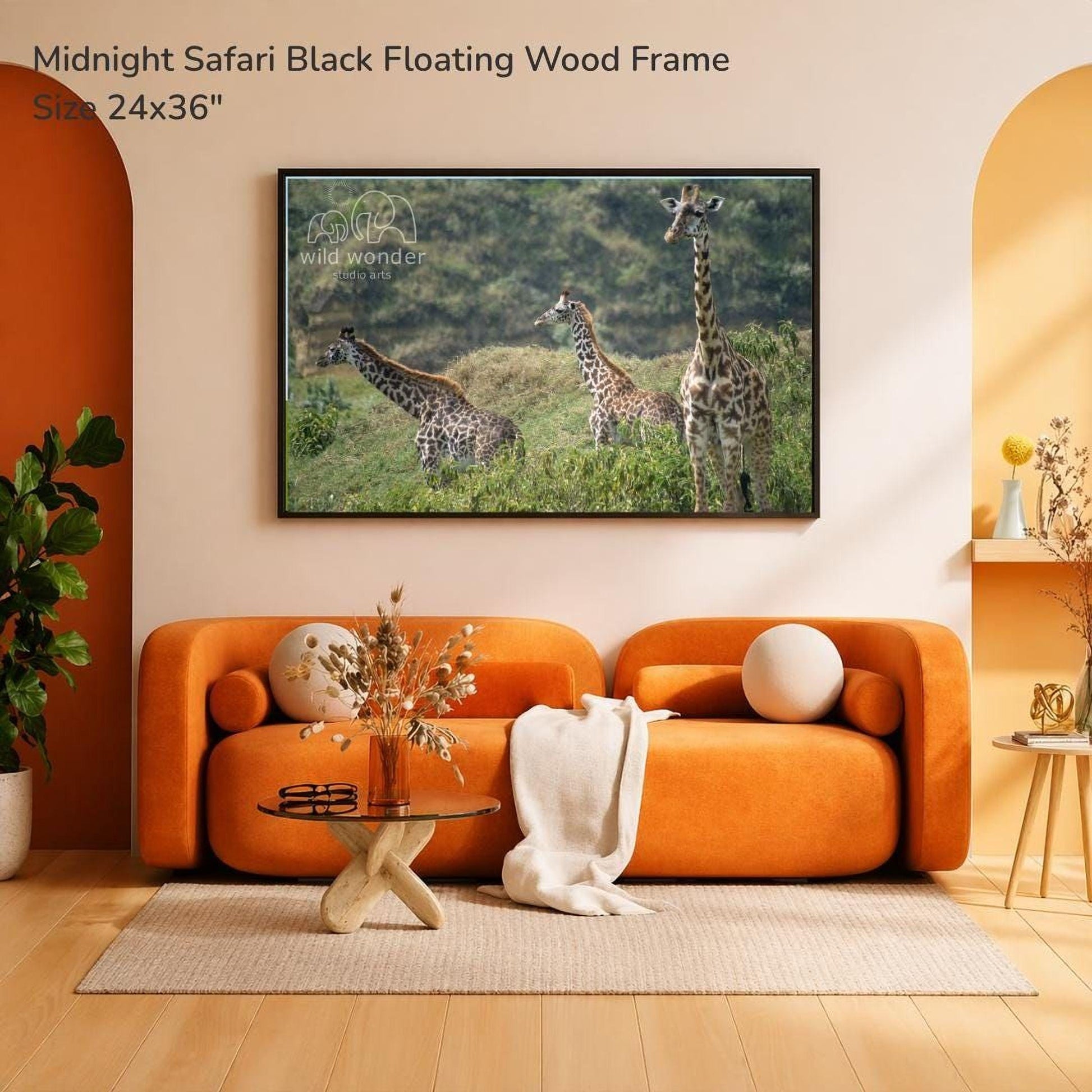 a large black floating wood frame hanging on a wall, depicting three giraffes in a grassy field. The frame is placed above a cozy orange couch, with a glass coffee table in front of it. The room has a warm, inviting atmosphere with a rug on the floor and some decorative elements like a potted plant and a vase.