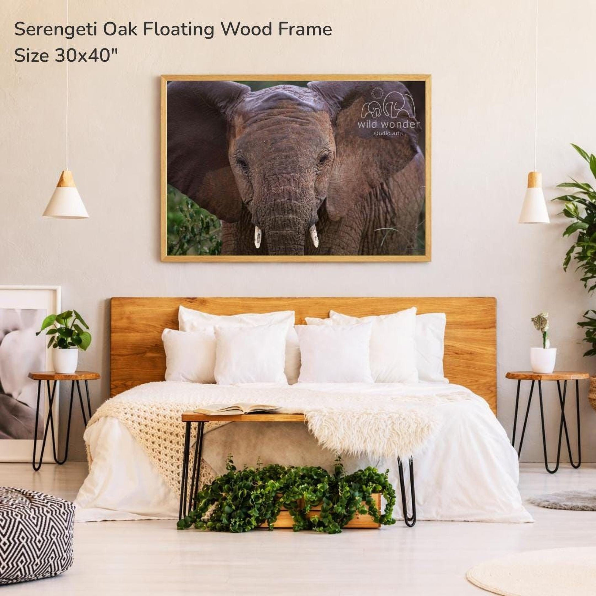 a serene bedroom setting with a large serene oak floating wood frame hanging above a bed with white bedding. The frame is framed in wood and has a size of 30x40 inches. The bed is adorned with white pillows and a white blanket, and there are two potted plants in the room. The overall atmosphere of the room is calm and peaceful, with a focus on the serene oak floating wood frame as the central decorative element.