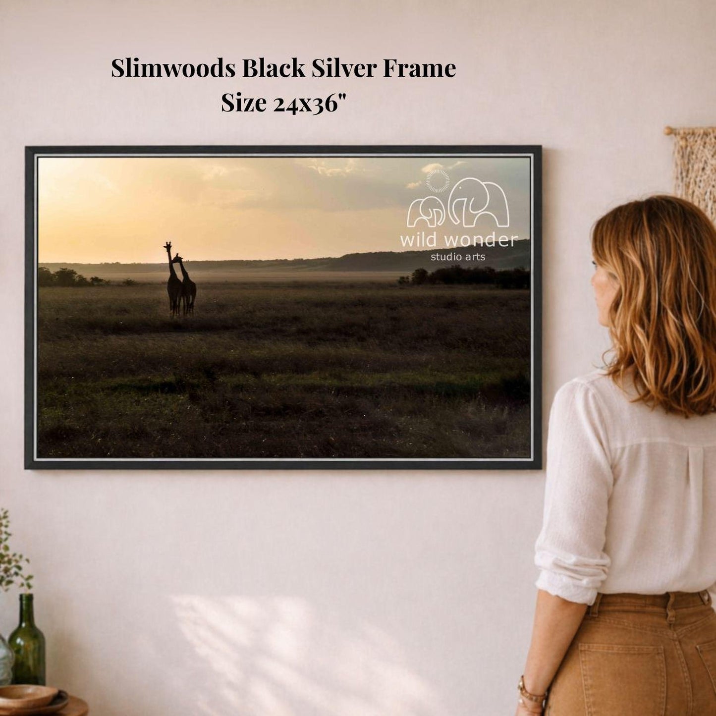 A woman is standing in front of a framed photograph of a giraffe in a grassy field, looking at the artwork.