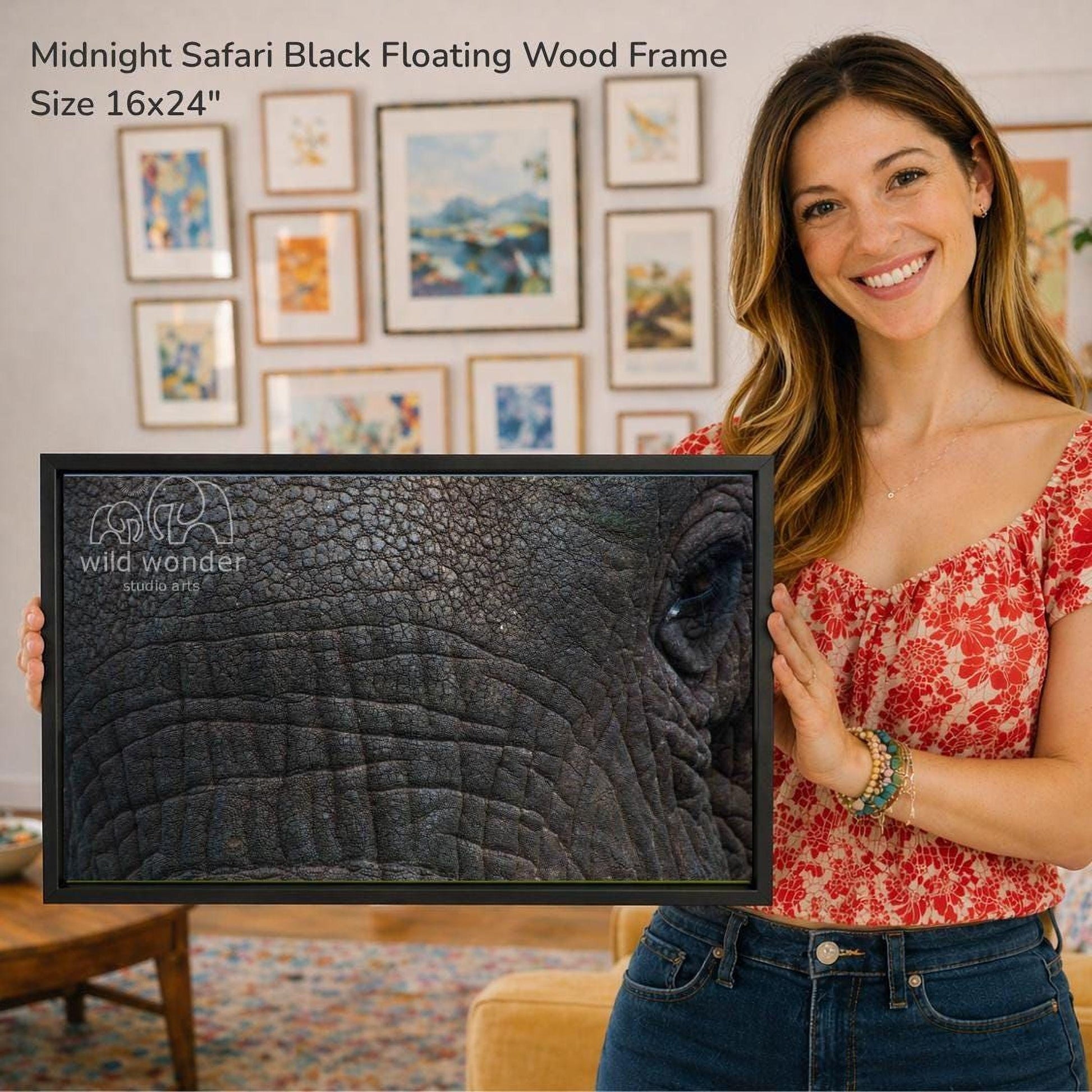 A woman is holding a framed photograph of an elephant's face, with a smiling expression. The background features a wall with various framed artworks.