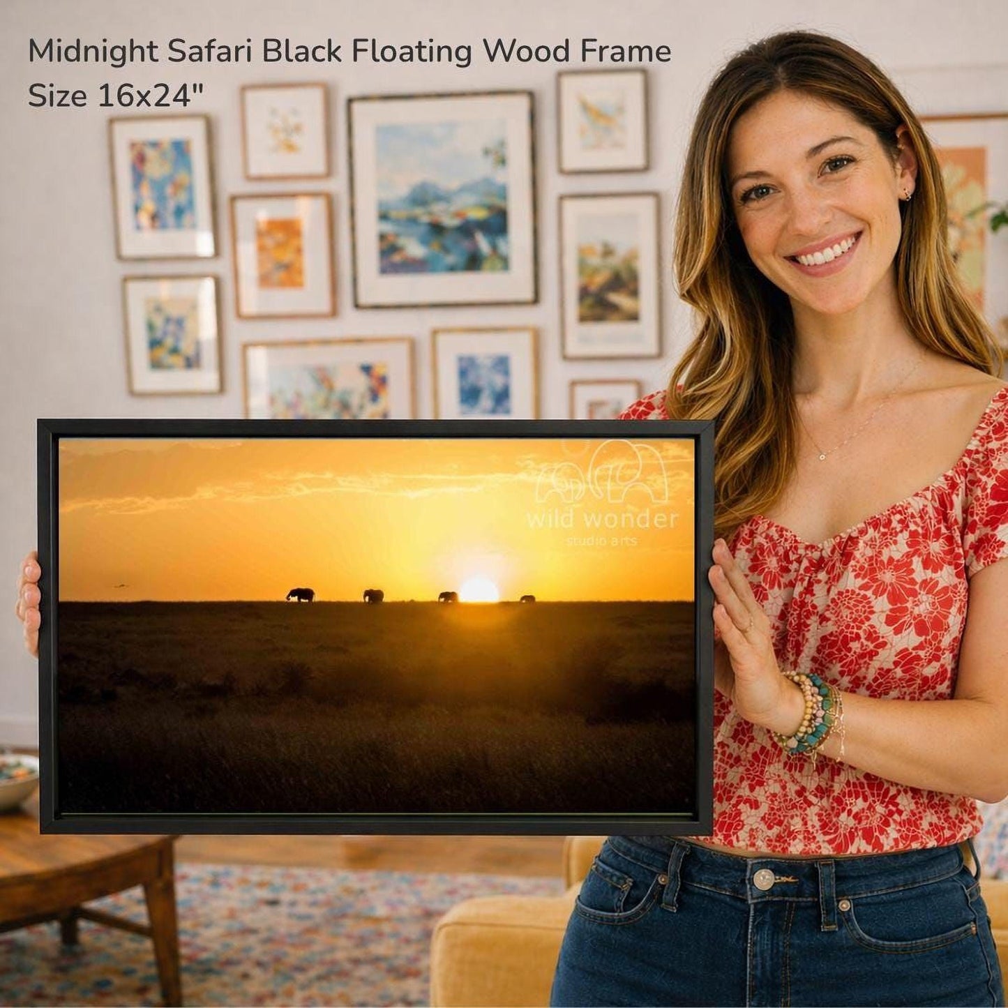 A woman is holding a framed picture of a sunset over a savannah, with a warm glow of the setting sun and silhouettes of elephants in the distance.