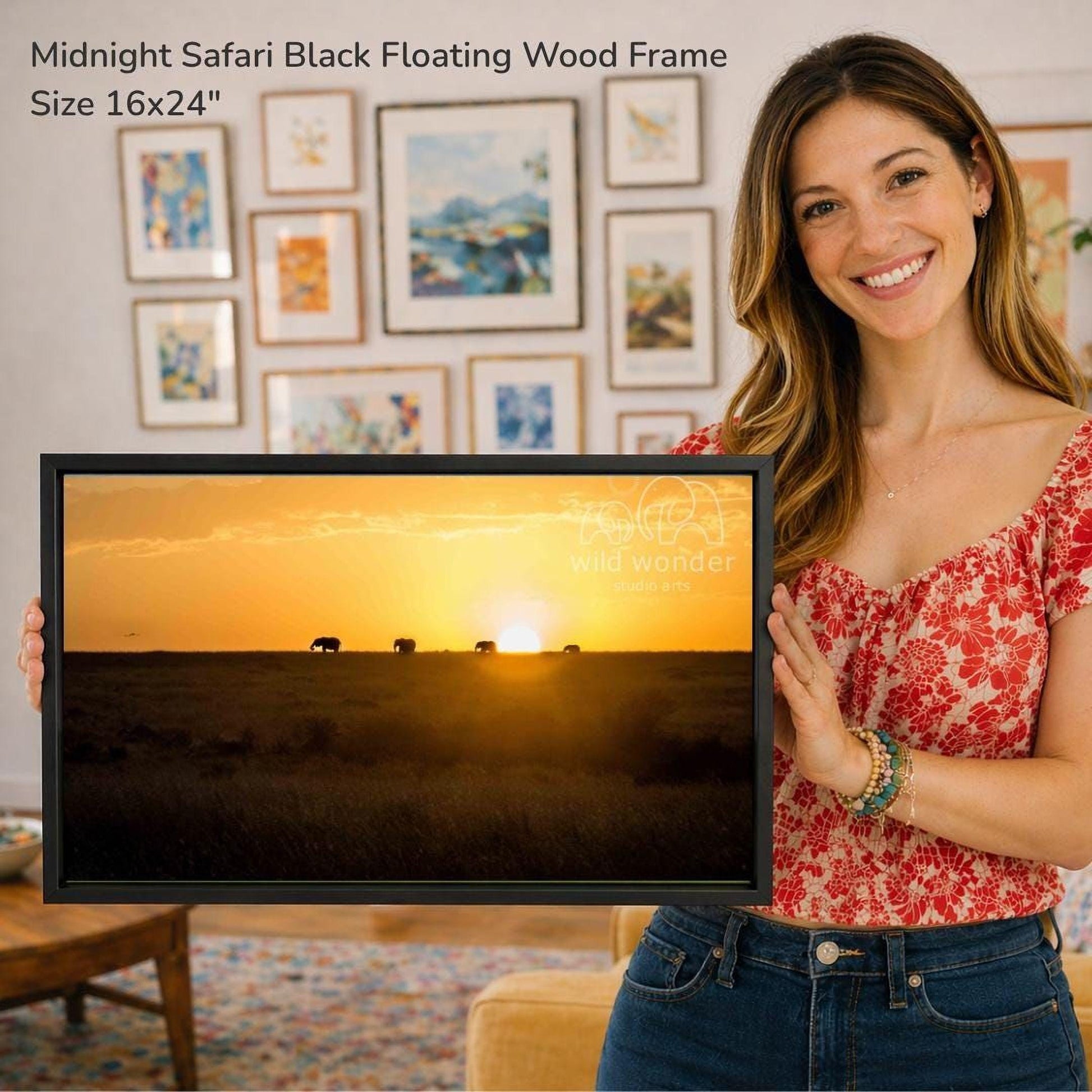 A woman is holding a framed picture of a sunset over a savannah, with a warm glow of the setting sun and silhouettes of elephants in the distance.