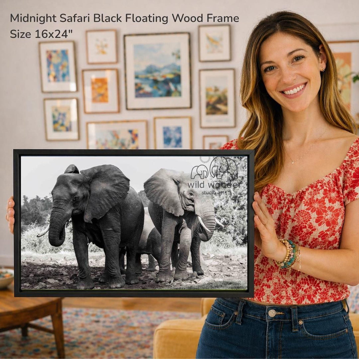 A woman is holding a framed photograph of three elephants in a natural setting, with a wall of framed artwork in the background.